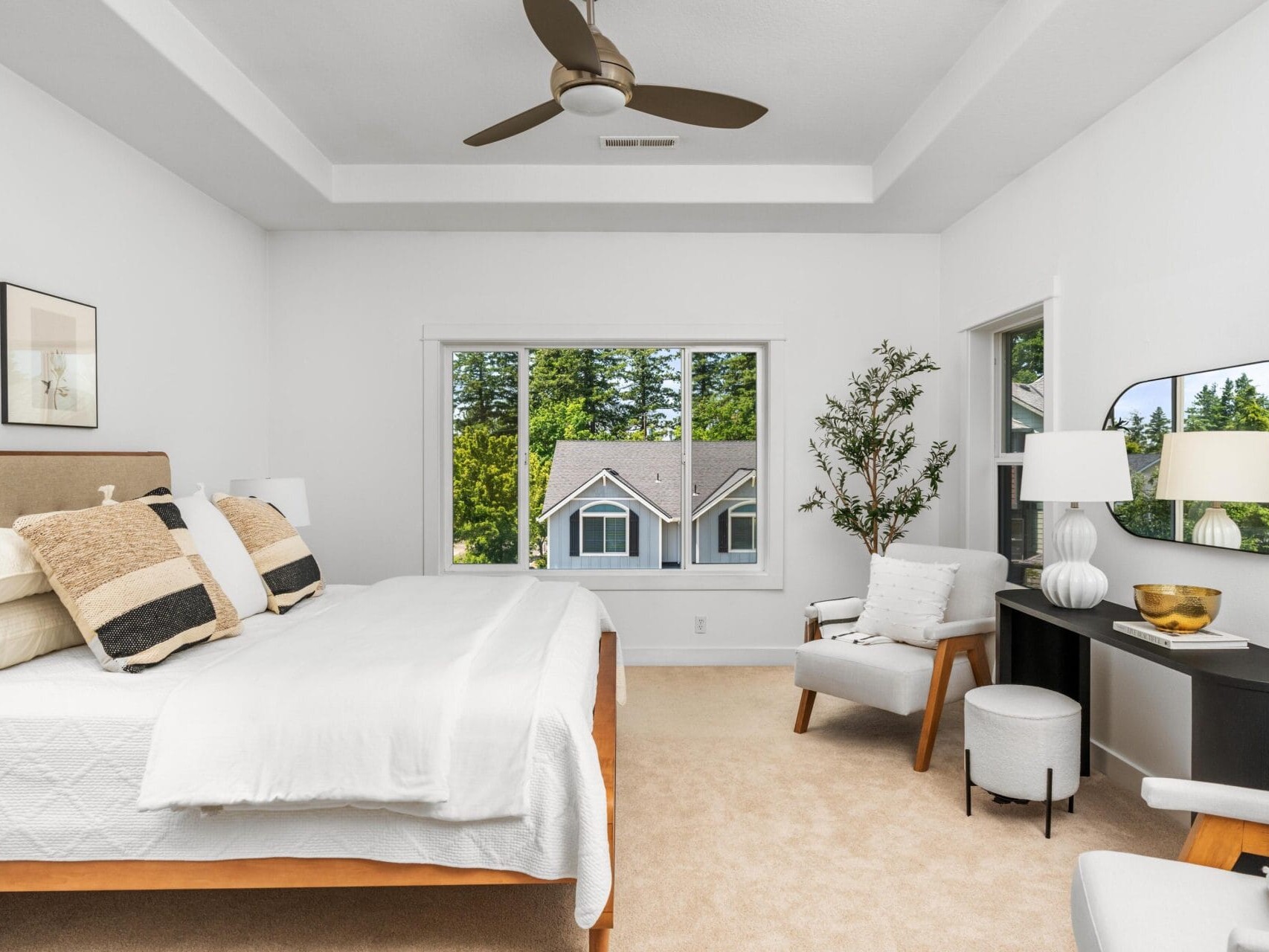 Modern bedroom with a large bed, neutral decor, and pillows; a window with a view of trees and rooftops; white armchair, black desk with a mirror, lamp, and plant; ceiling fan overhead.