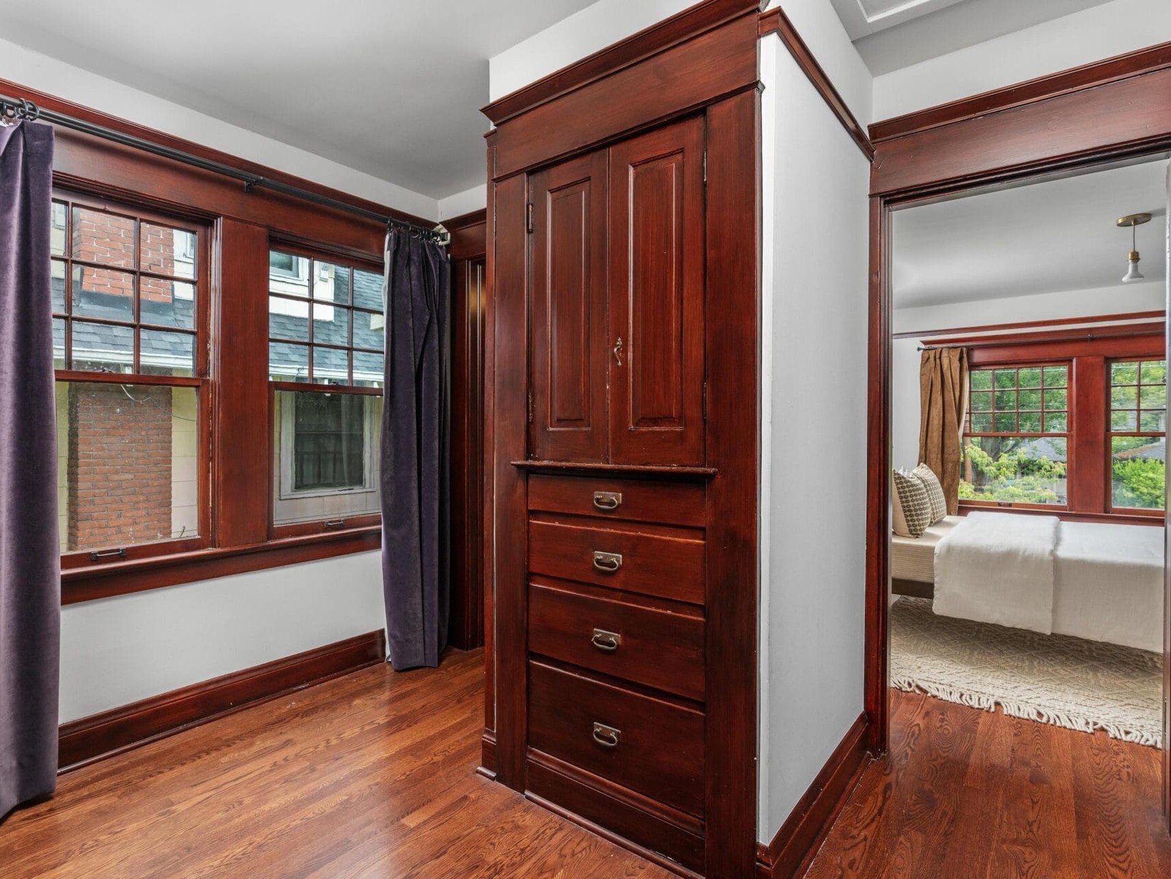 A hallway with rich dark wood trim, built-in drawers and a cabinet, hardwood floors, and large windows with dark curtains. An adjoining bedroom with a bed and windows is visible.