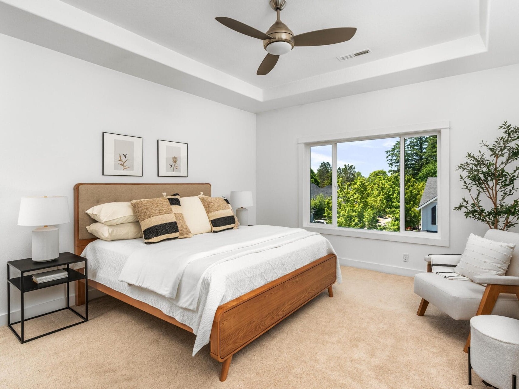 Modern bedroom with a wooden bed, white bedding, neutral pillows, two black nightstands with lamps, a large window with a view of greenery, framed art on the wall, a ceiling fan, and an armchair with a blanket.
