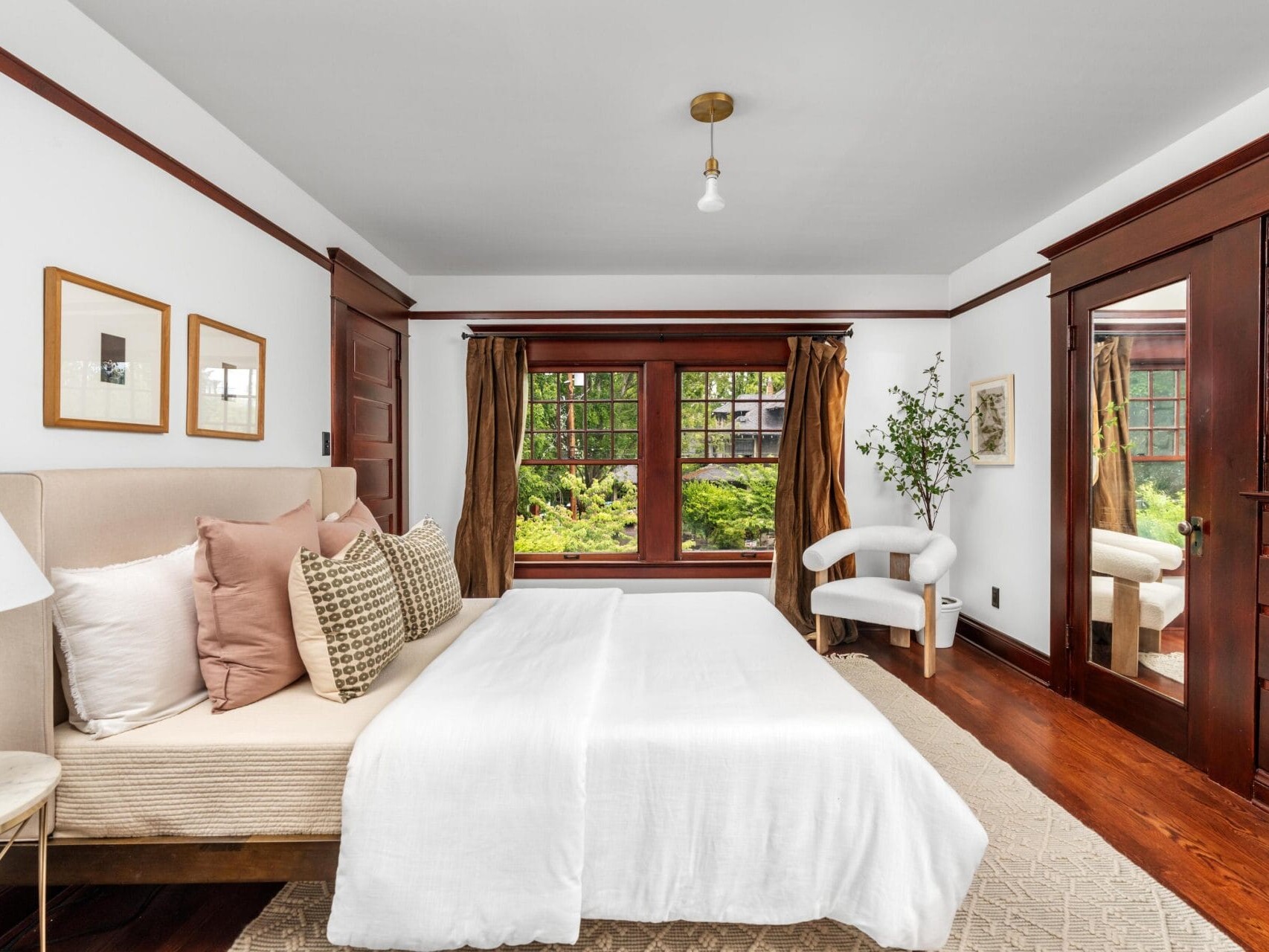 A cozy bedroom with a beige bed, neutral pillows, white bedding, dark wood trim, large windows with brown curtains, a white accent chair, a green plant, and a mirrored wardrobe. Natural light fills the room.