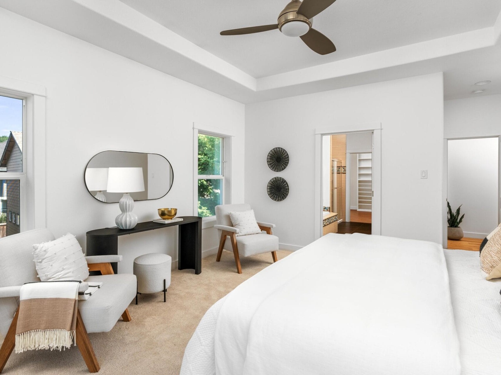 A bright, modern bedroom with white walls and bedding, a ceiling fan, a bed with patterned pillows, two armchairs, a black desk with a lamp and mirror, and a view into a bathroom. Large windows let in natural light.