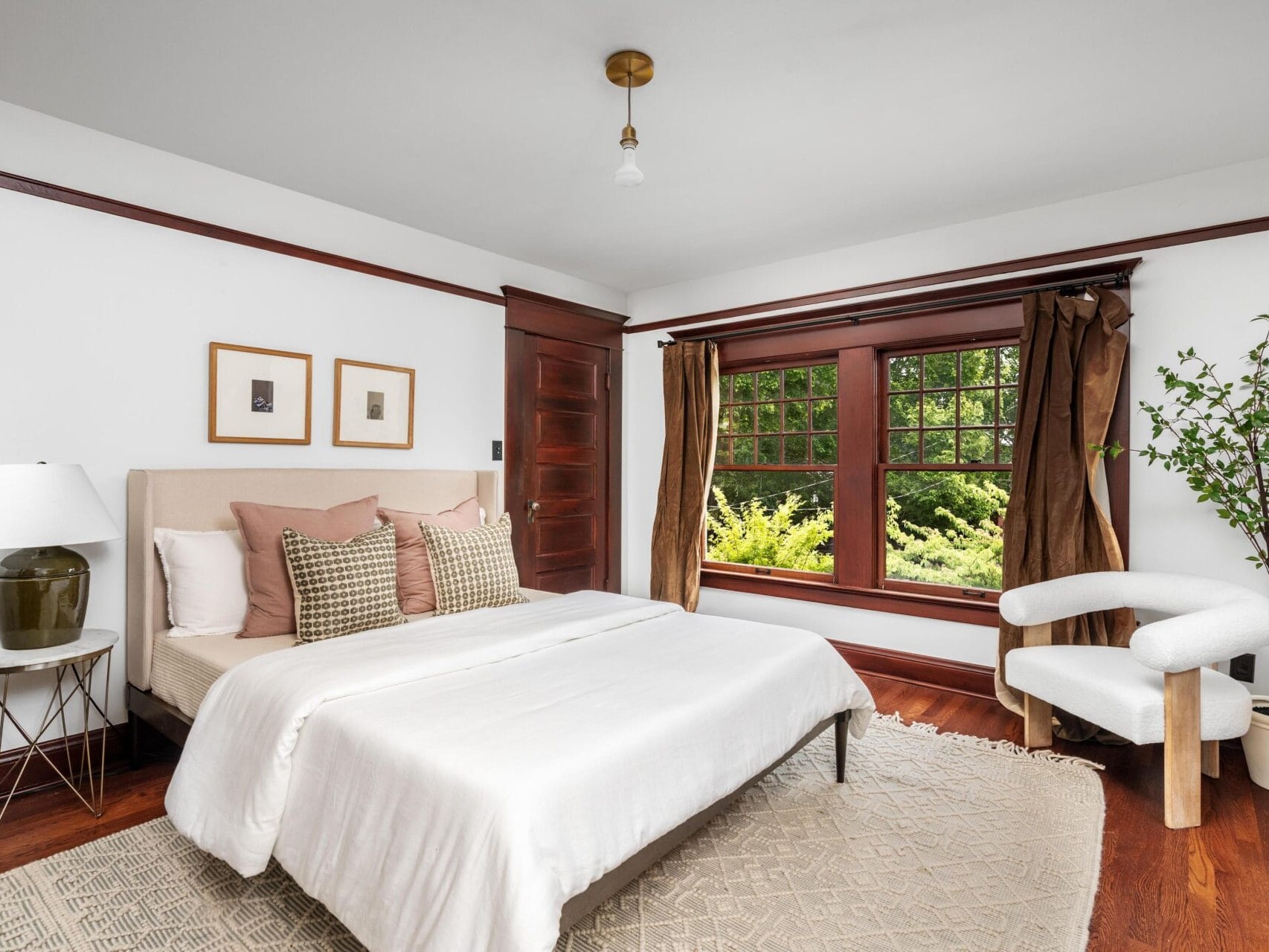 A bright, cozy bedroom with a beige bed, neutral bedding, accent pillows, a white rug, and a side table with a lamp. Large window with brown curtains overlooks greenery; a modern chair and potted plant sit nearby.
