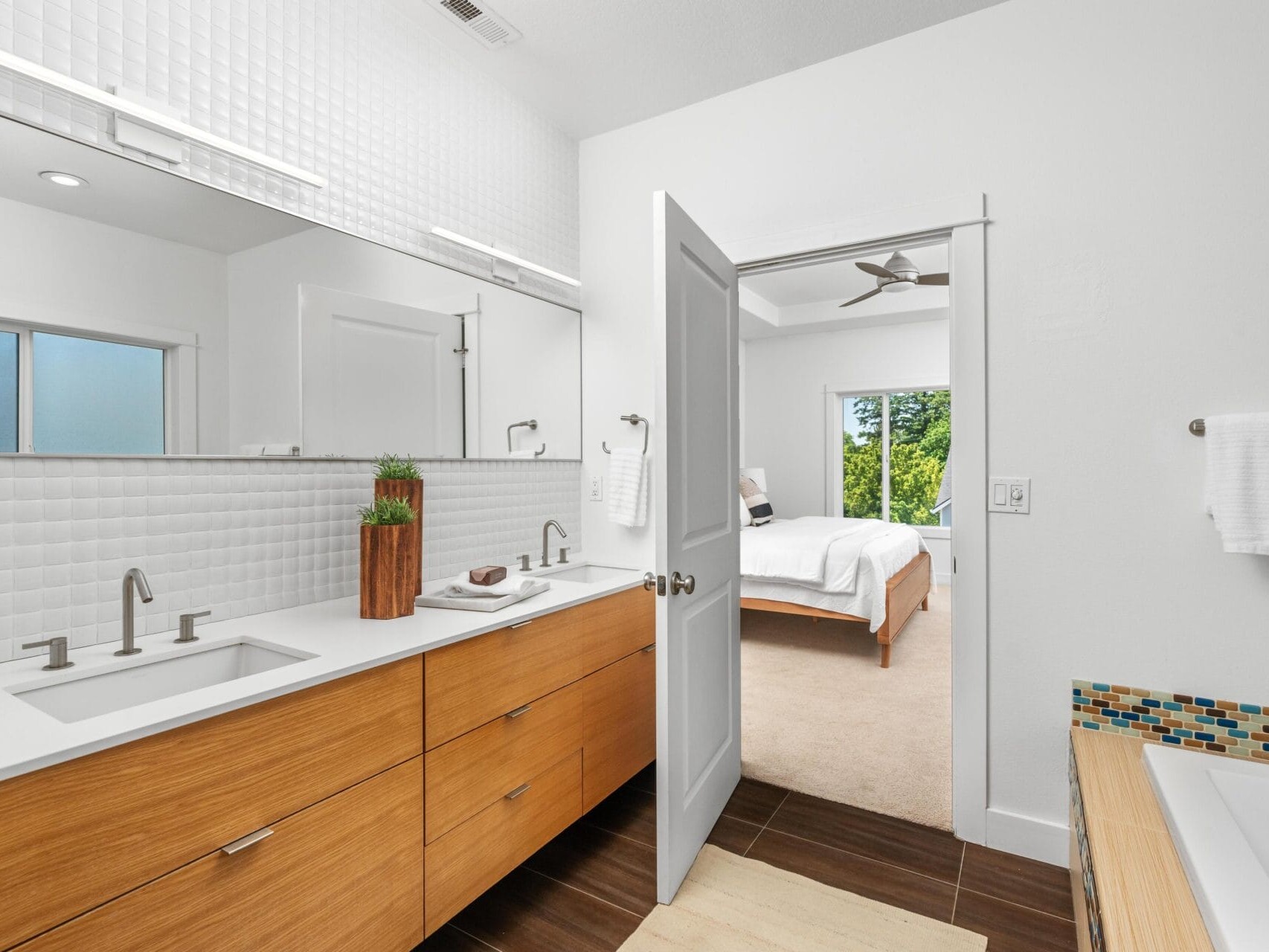 Modern bathroom with double sinks, wood cabinets, and a large mirror; a door leads to a bright bedroom with a bed, ceiling fan, and large windows overlooking greenery.