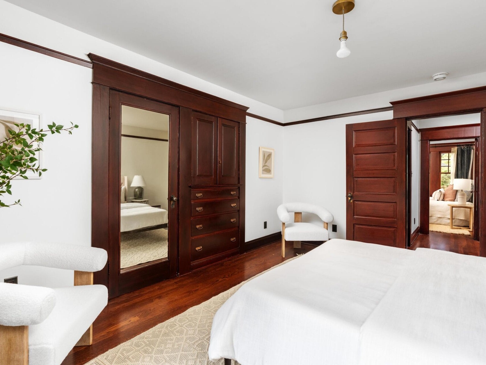 A bright bedroom with white walls, dark wood trim and doors, a bed with white bedding, two white armchairs, a large built-in wardrobe with a mirror, and minimal decor including framed art and a leafy plant.