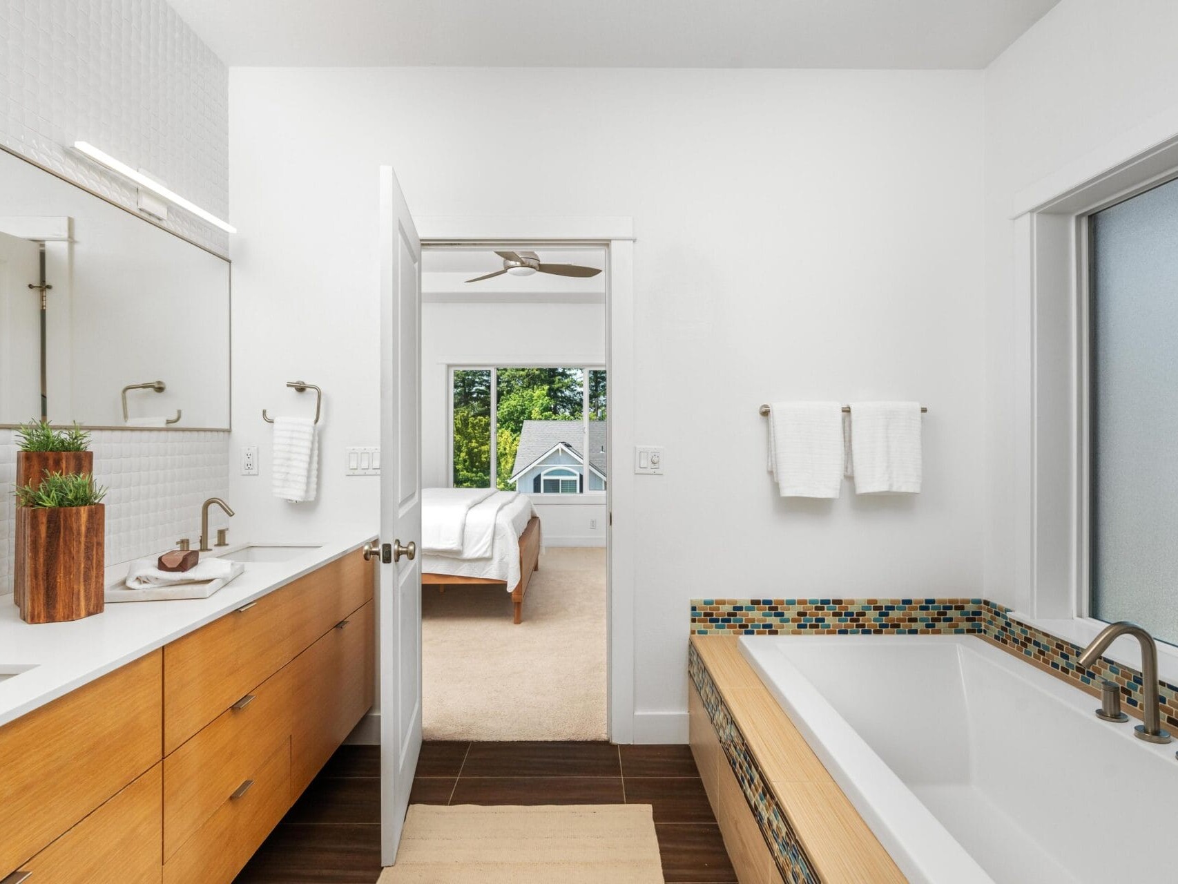 Modern bathroom with wooden cabinets, a large vanity mirror, double sinks, and a bathtub with mosaic tile accents. A door opens to a bedroom with a bed and large windows showing greenery outside. Towels hang by the bathtub.