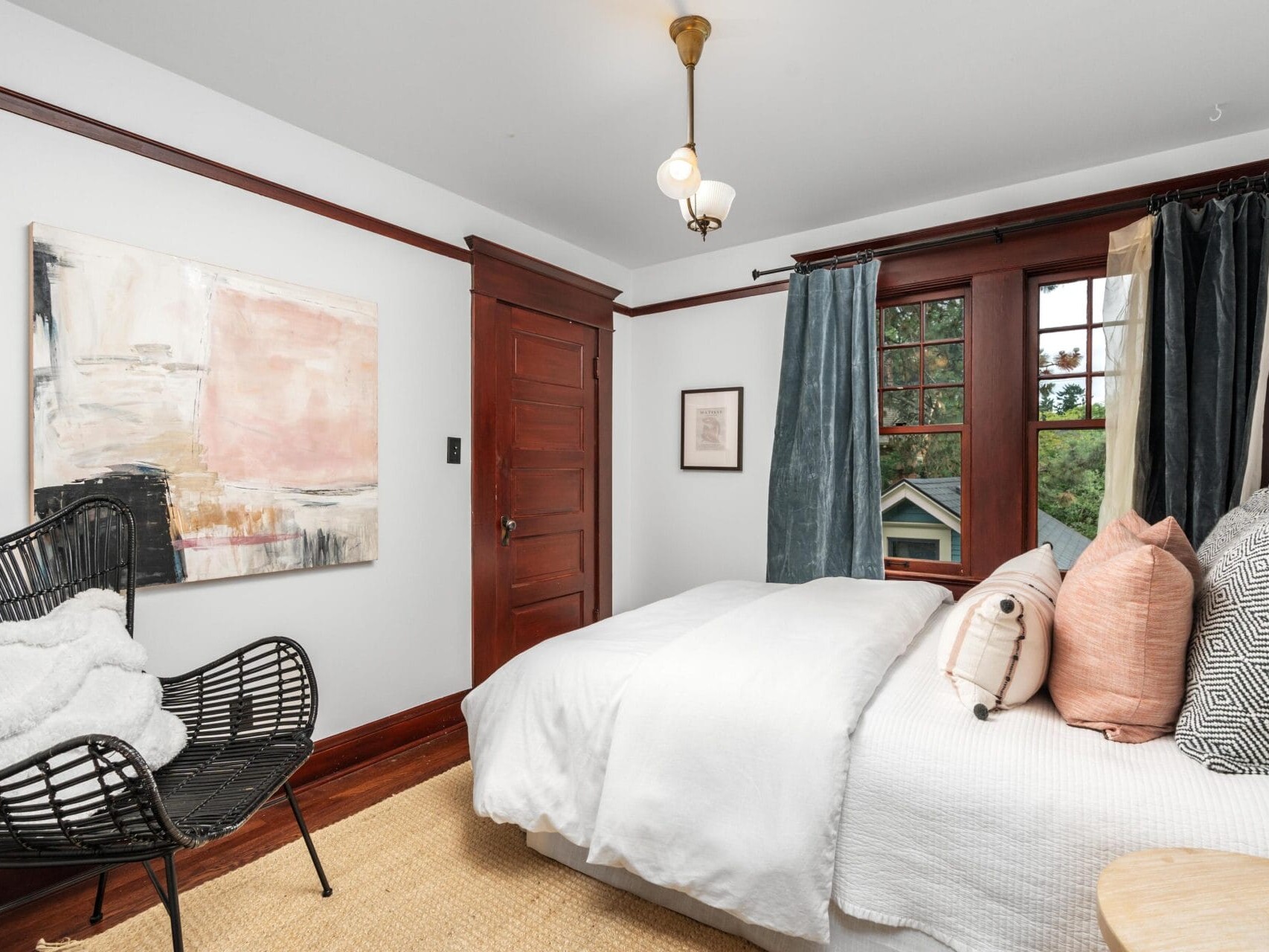 Bright bedroom with white bedding, pink and gray pillows, a black wicker chair with a white throw, abstract wall art, dark wood trim, blue curtains, and a window overlooking greenery.