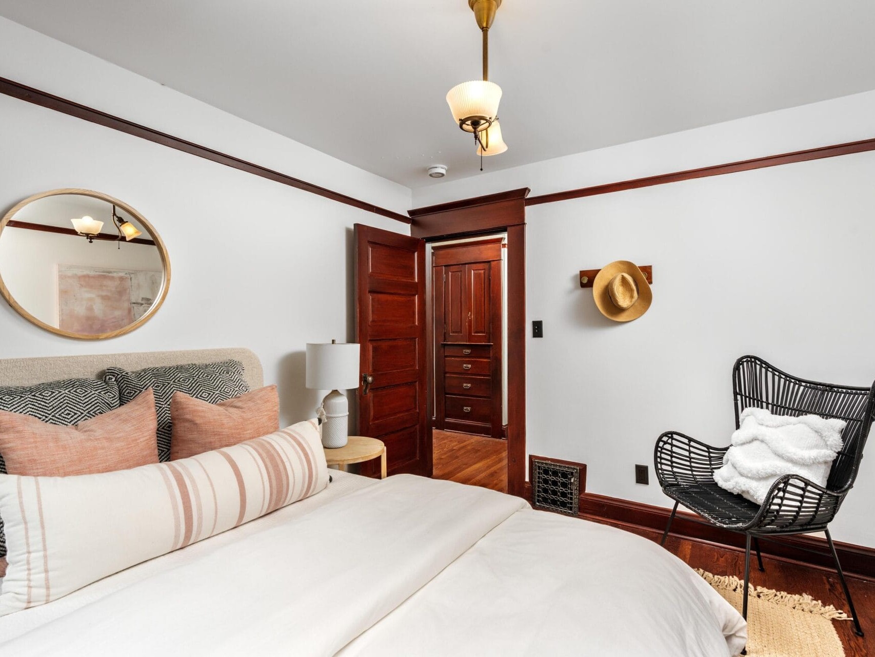 A cozy bedroom with a neatly made bed, textured pillows, a round mirror above the headboard, a black wicker chair with a white blanket, and a wooden door with a hat hanging on the wall beside it.