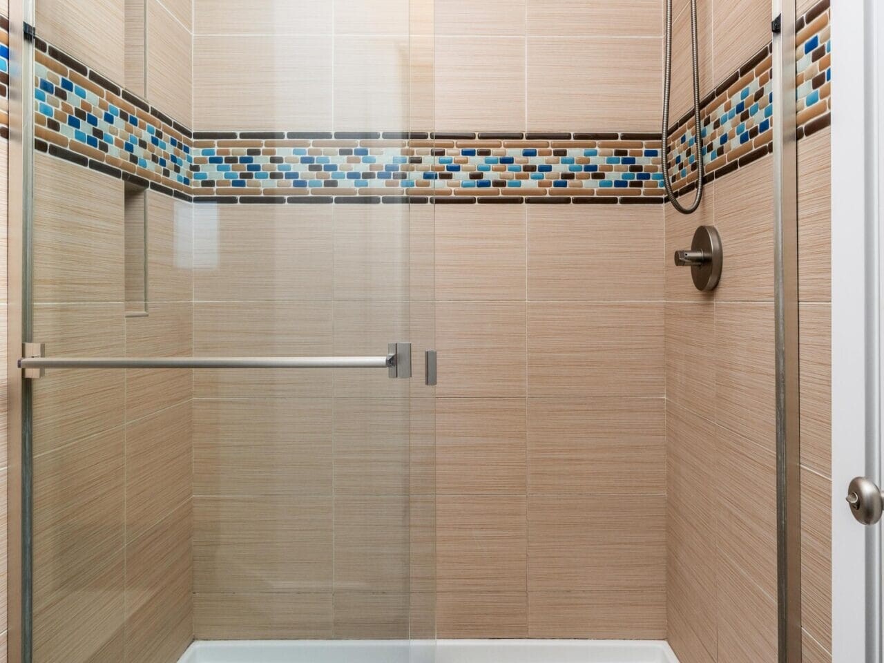 A modern shower with sliding glass doors, beige tiles, a horizontal strip of multicolored mosaic tiles, a built-in shelf, a wall-mounted showerhead, and a metal handle.
