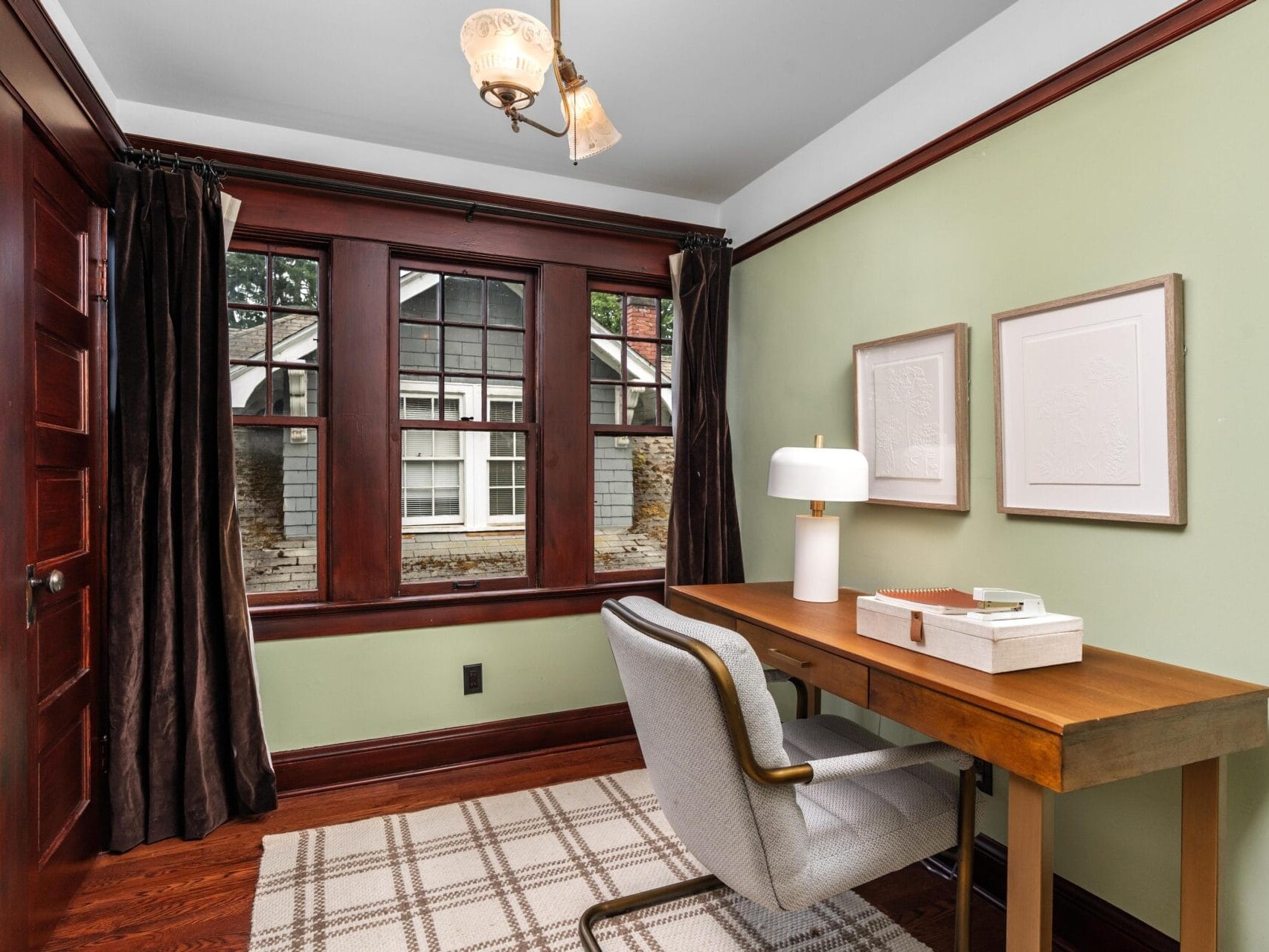 A cozy home office with green walls, wooden trim, a large window, a wooden desk with a lamp and tray, a gray upholstered chair, plaid rug, and two framed artworks on the wall.