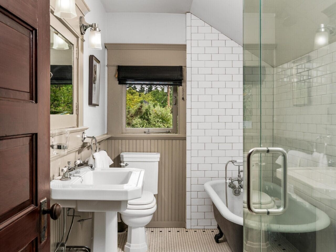 A bright bathroom with white subway tile walls, hexagonal floor tiles, a pedestal sink, a toilet, a black clawfoot tub with a shower, a wooden door, and a window with a black shade.