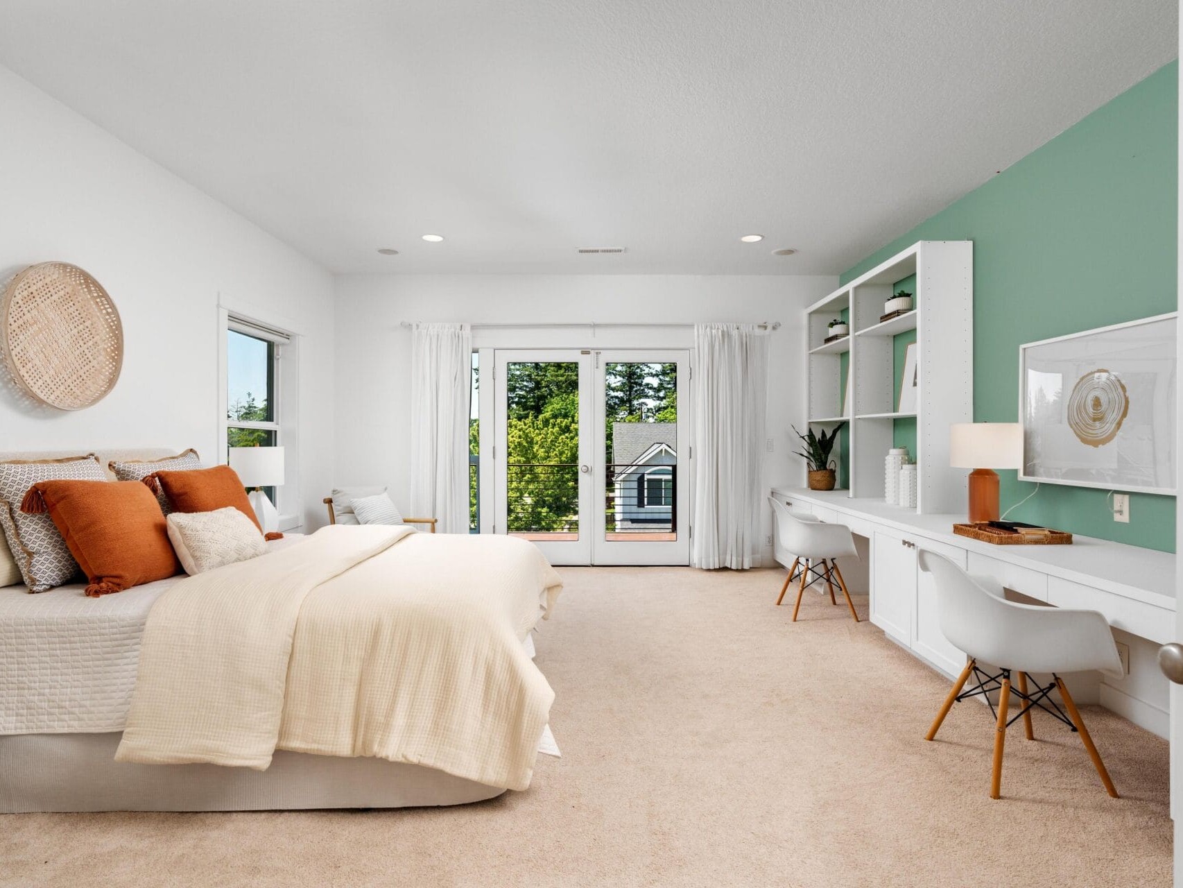 Modern bedroom with a neatly made bed, neutral bedding, and orange accent pillows. There’s a built-in desk with shelves, two chairs, and a mint green accent wall. French doors open to a balcony with a view of trees.