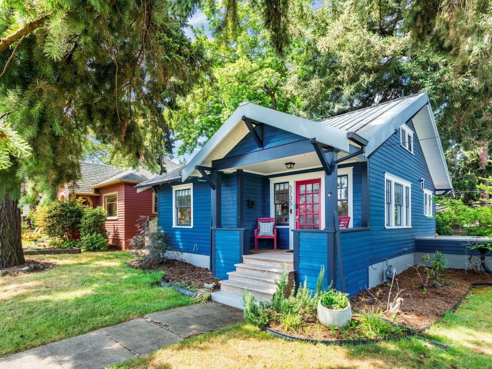 Blue cottage-style house with a covered front porch, white trim, two red chairs, and surrounded by green grass, trees, and shrubs on a sunny day. A neighboring house is partially visible to the left.