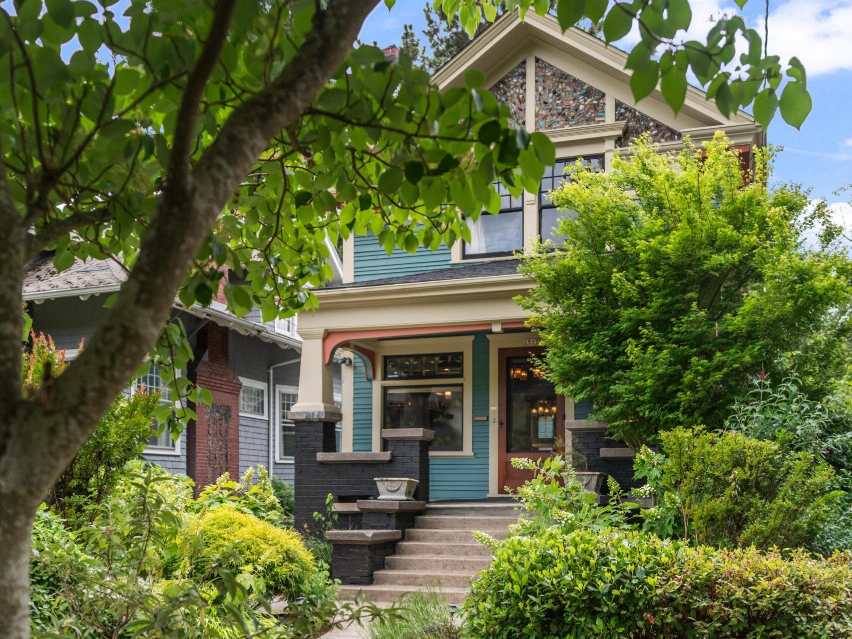 A charming two-story house with teal siding, ornate trim, and a covered porch, surrounded by lush green trees and shrubs on a sunny day.