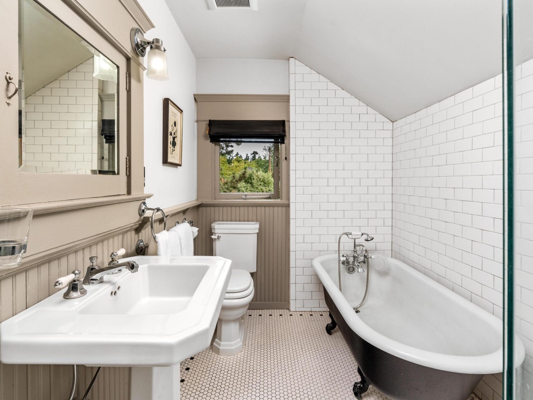 A bright bathroom with a pedestal sink, toilet, clawfoot bathtub, white subway tile walls, beadboard wainscoting, a wall mirror, and a window with a view of greenery outside.