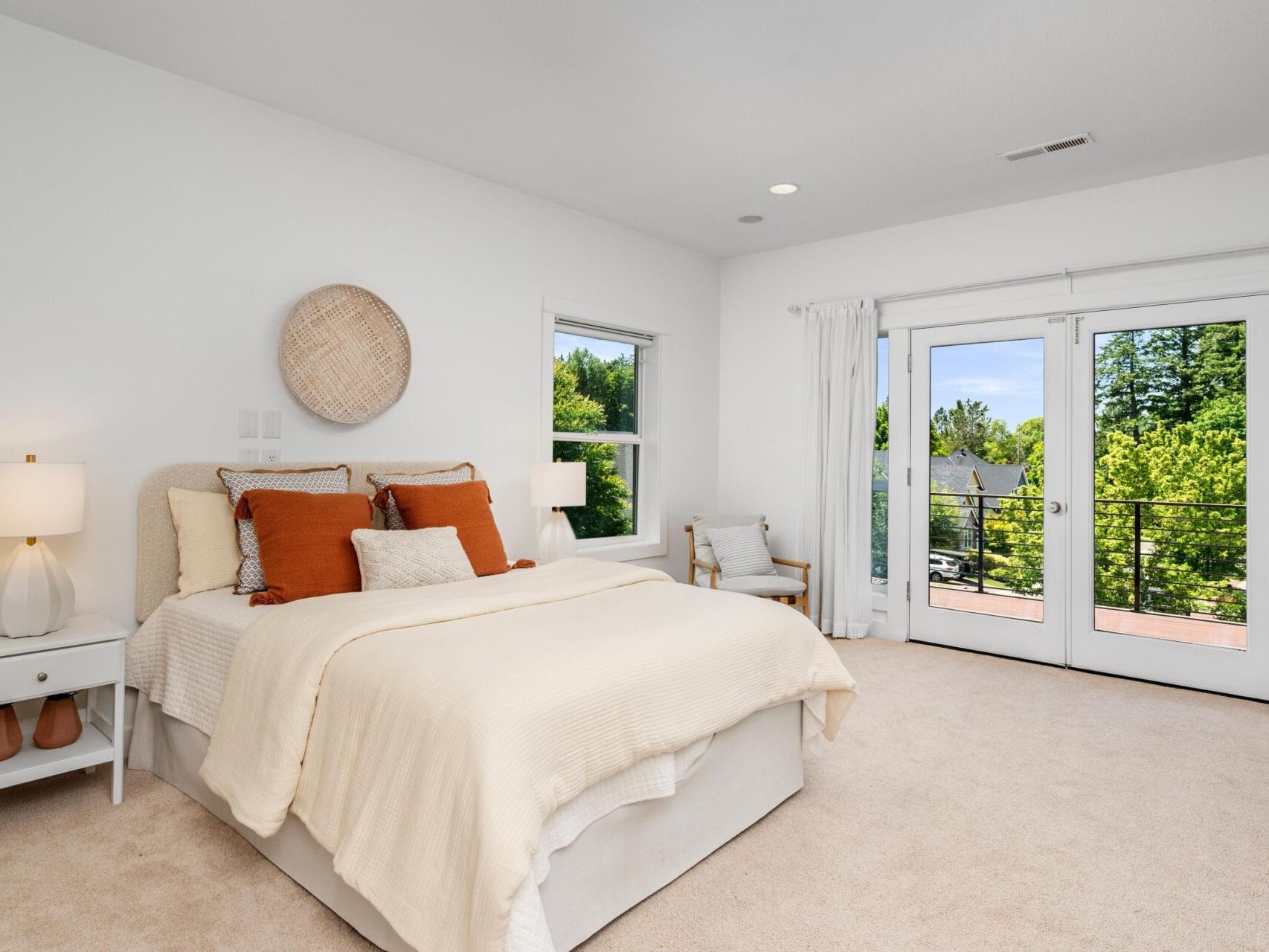 Bright bedroom with a neatly made bed featuring orange and beige pillows, a nightstand with a lamp, a chair, and large windows with French doors opening to a balcony overlooking greenery.