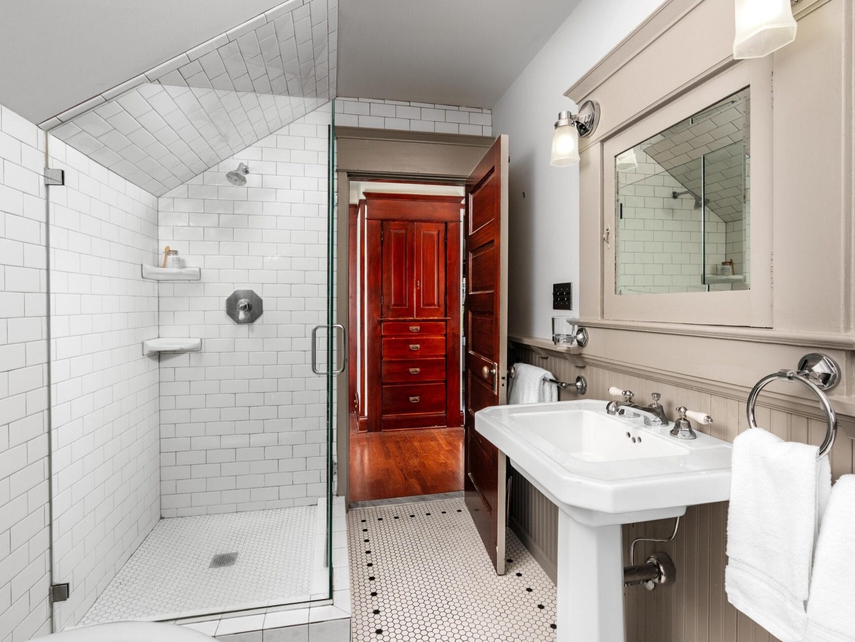 A bright bathroom with white subway tile walls, glass shower enclosure, pedestal sink, and a large mirror. Two white towels hang on a rack. An open wooden door leads to a hallway with hardwood floors.