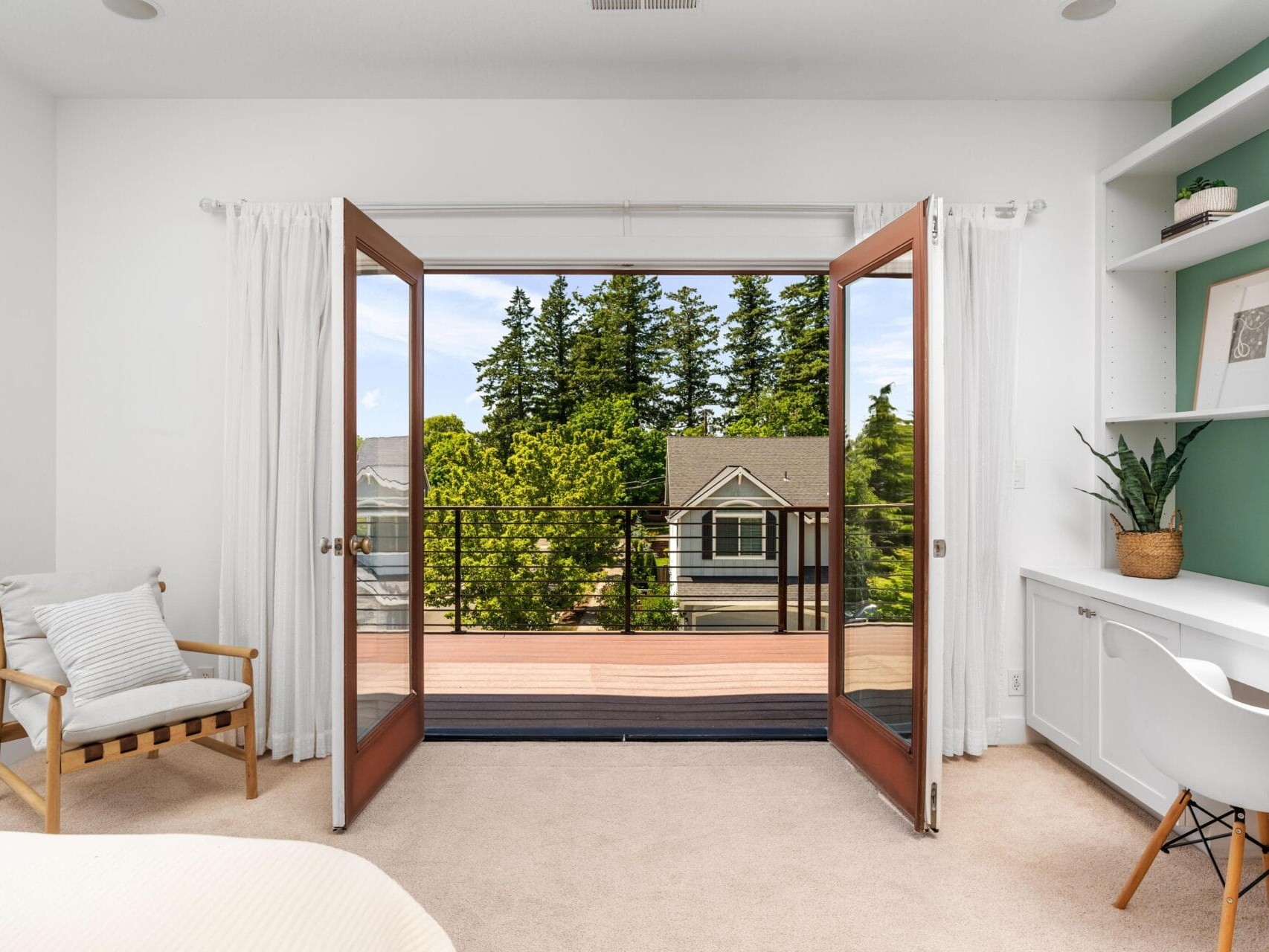 Bright bedroom with open double doors leading to a balcony overlooking trees and neighboring houses; features a cozy chair, built-in desk, shelves, and white curtains.