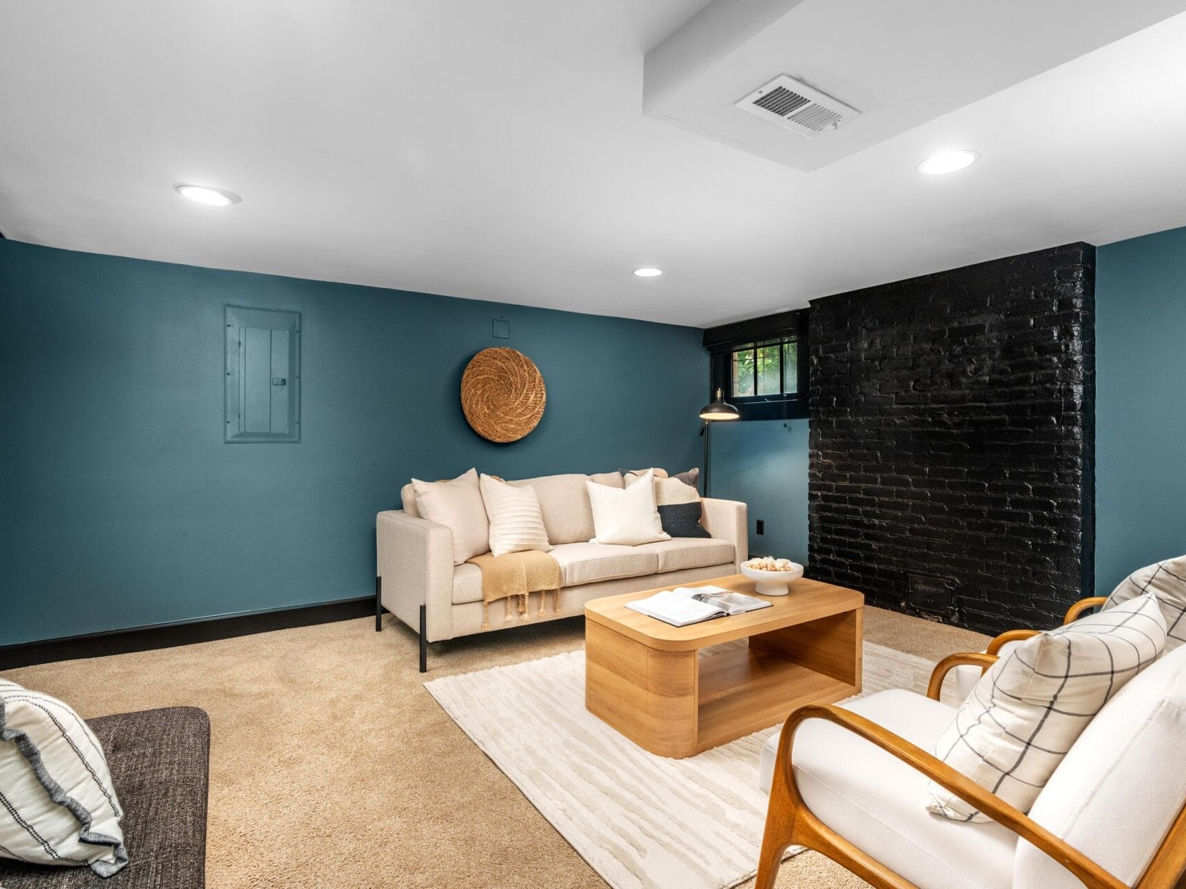 A cozy living room with blue walls, a black painted brick accent wall, beige carpet, a white sofa, a wooden chair with cushions, a wooden coffee table, and a woven wall decoration.