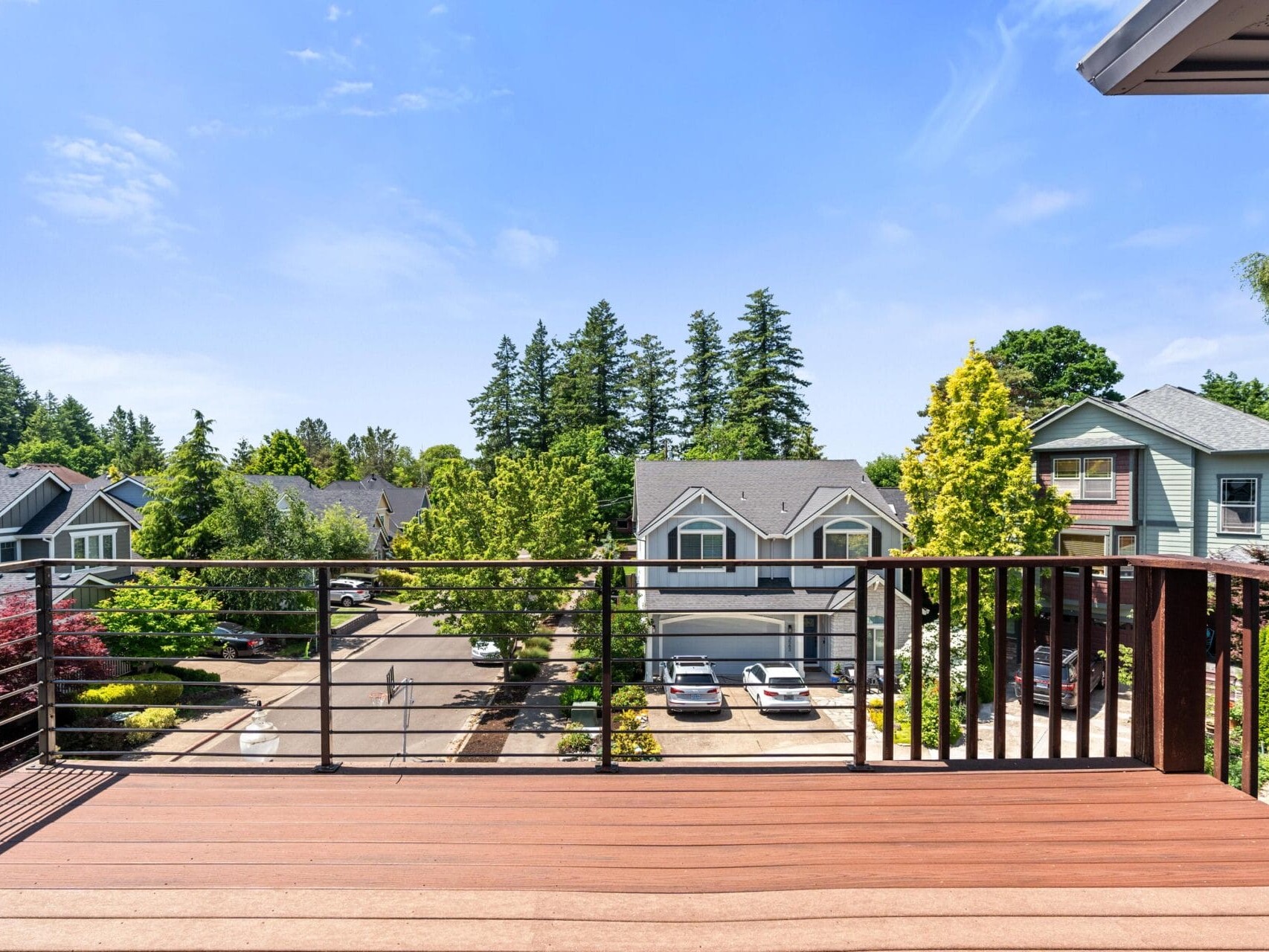 A wooden balcony with metal railings overlooks a quiet suburban street lined with trees and houses under a bright blue sky.