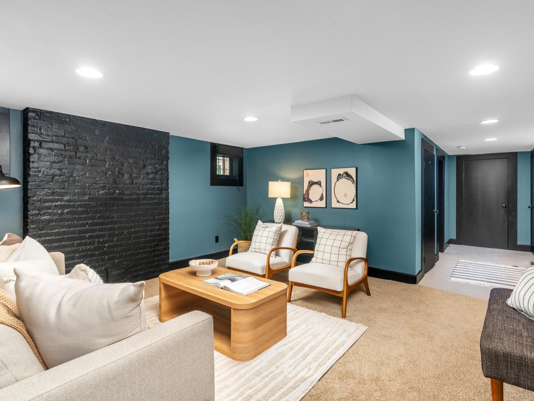 A modern basement living room with teal walls, a black brick accent wall, beige carpet, light-colored sofas and chairs, a wooden coffee table, wall art, and a hallway with a laundry area in the background.