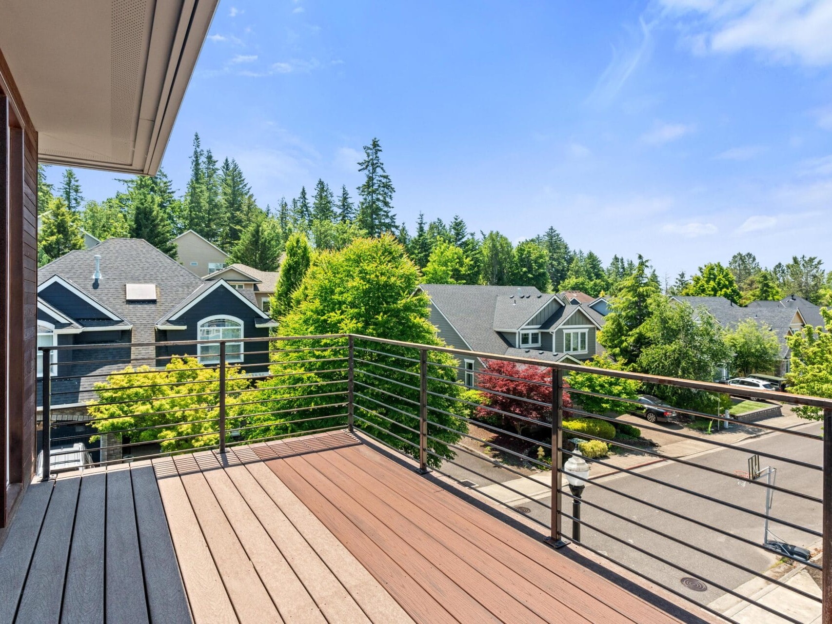 View from a balcony with wooden flooring and metal railing, overlooking a suburban street lined with trees and houses under a bright blue sky with scattered clouds.