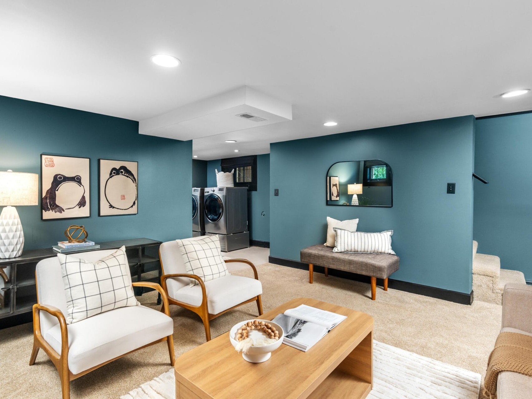 A modern basement living area with blue-green walls, beige carpet, and white ceiling. It features two armchairs, a wooden coffee table, a sofa, wall art, a bench, a mirror, and a laundry area in the background.