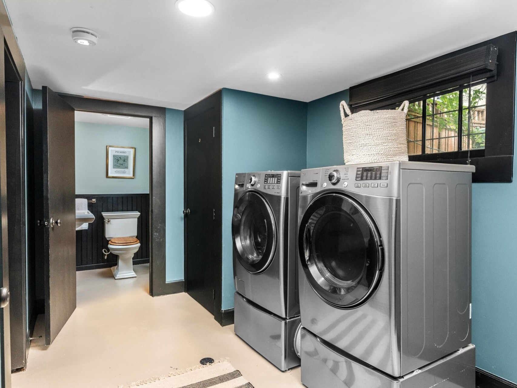 Modern laundry room with blue walls, featuring a front-loading washer and dryer, a woven basket on top, black doors, and a window. A bathroom with a toilet is visible through an open doorway.