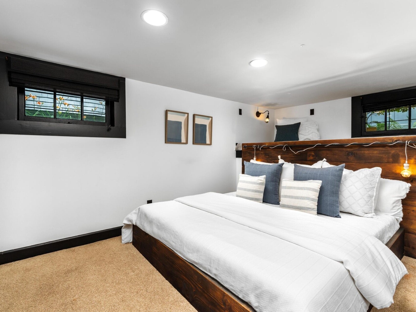 A modern bedroom with a large wooden bed, white bedding, and multiple pillows. The room features two small windows with stained glass, beige carpet, and minimal wall decor, creating a clean and cozy atmosphere.