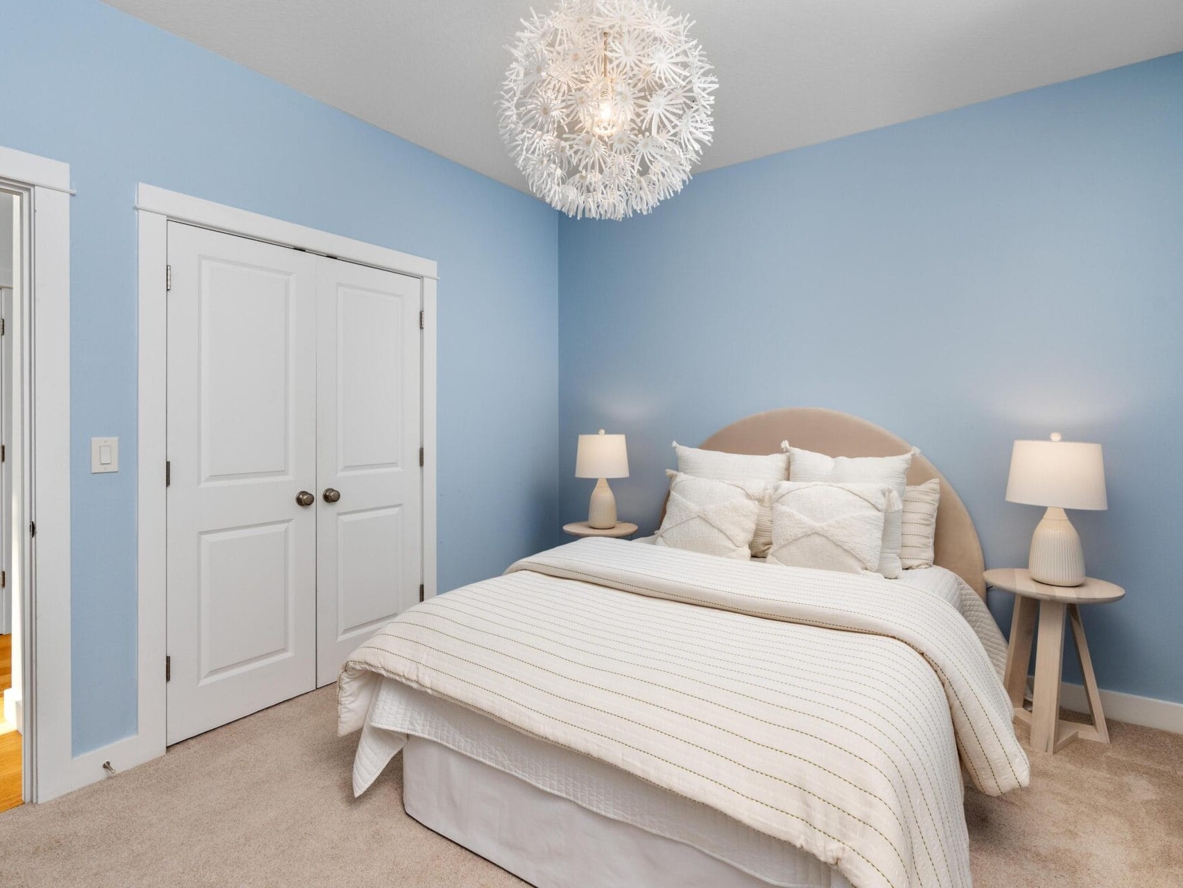 A cozy bedroom with light blue walls, a double bed with white bedding and pillows, two round bedside tables with lamps, a modern white chandelier, and a closed double-door closet.