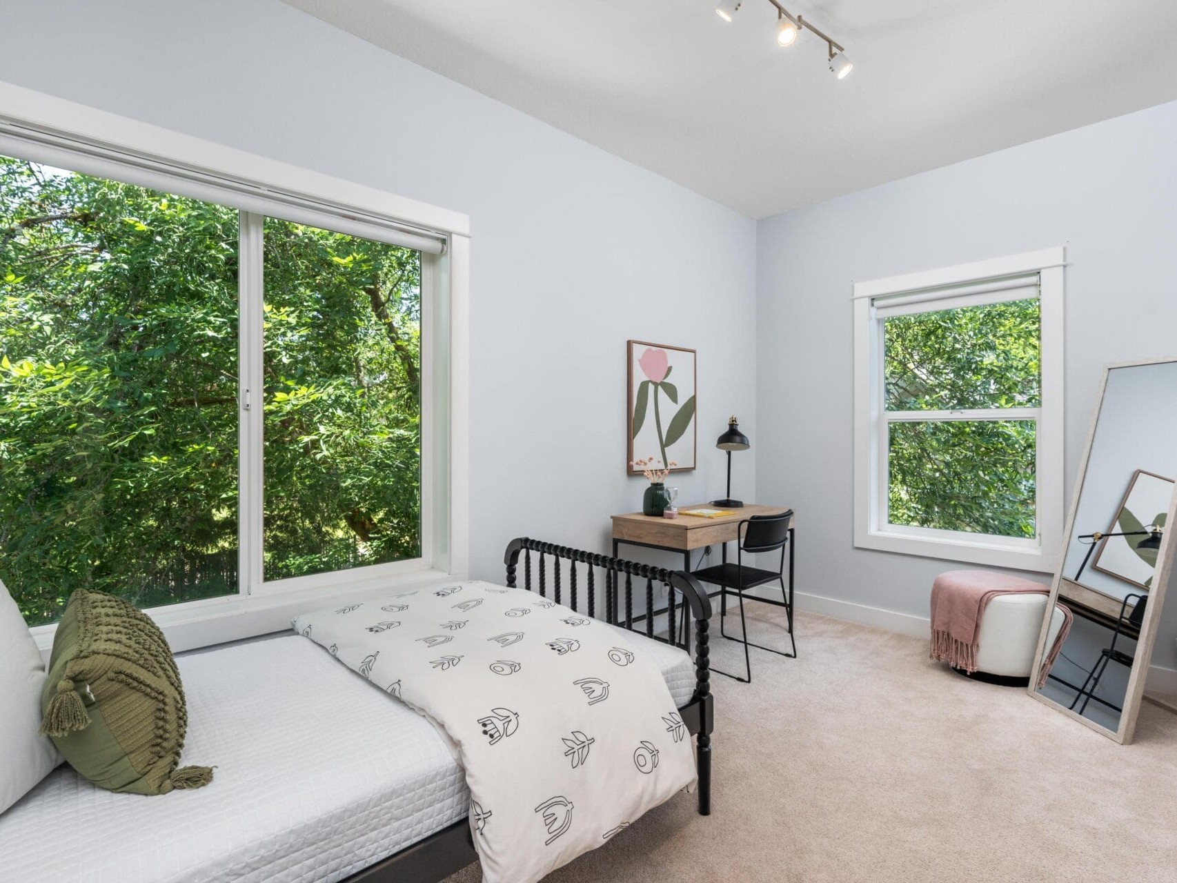 A bright, minimalist bedroom with a single bed, green pillow, desk and chair, framed artwork, full-length mirror, and large windows showing leafy trees outside. The walls are light blue and the carpet is beige.