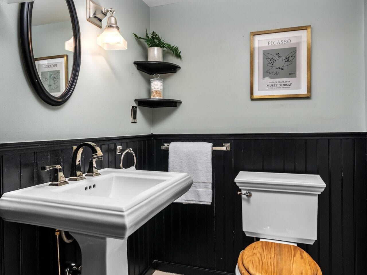 A small bathroom with pale green walls and black wainscoting features a pedestal sink, a toilet with a wooden seat, an oval mirror, wall shelves with a plant, a towel bar, and framed art on the wall.