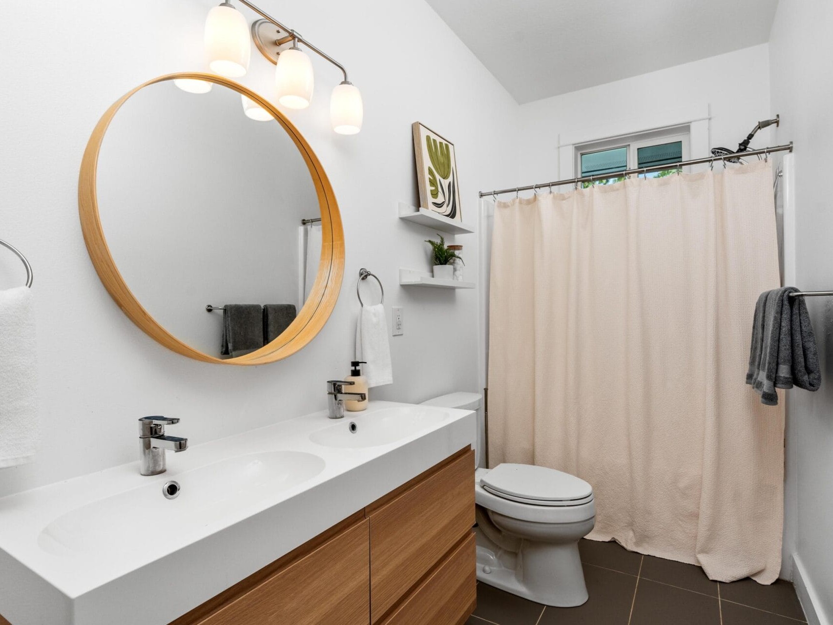 Modern bathroom with a double sink vanity, round mirror, wall-mounted lights, white walls, towel racks, a toilet, beige shower curtain, floating shelves with decor, and a small window above the shower.