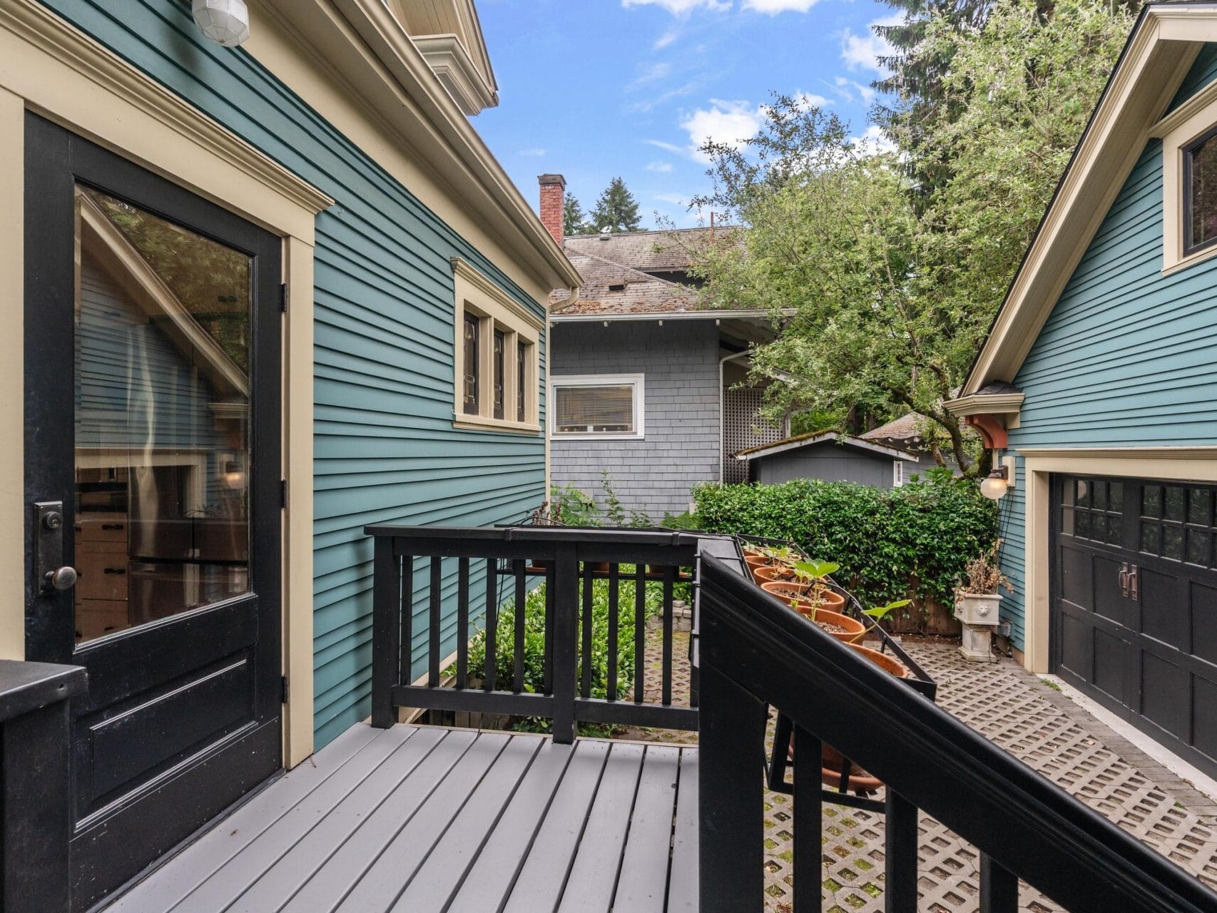 A small gray deck with black railing leads to a backyard between two blue houses with cream trim. Potted plants line the patio and a black garage door is visible on the right. Trees and greenery surround the area.