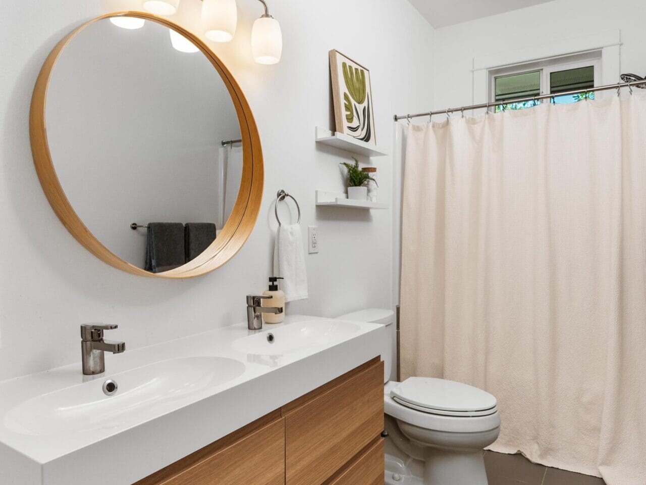 Modern bathroom with a double-sink wooden vanity, round mirror, wall-mounted lights, shelves with decor, white toilet, beige shower curtain, and gray tile floor. The walls are white and the space is bright and clean.