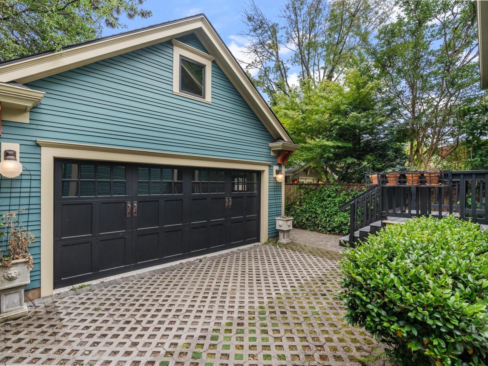 A teal detached garage with black double doors sits beside a paved driveway. To the right, a wooden deck with potted plants is surrounded by lush green trees and shrubs.