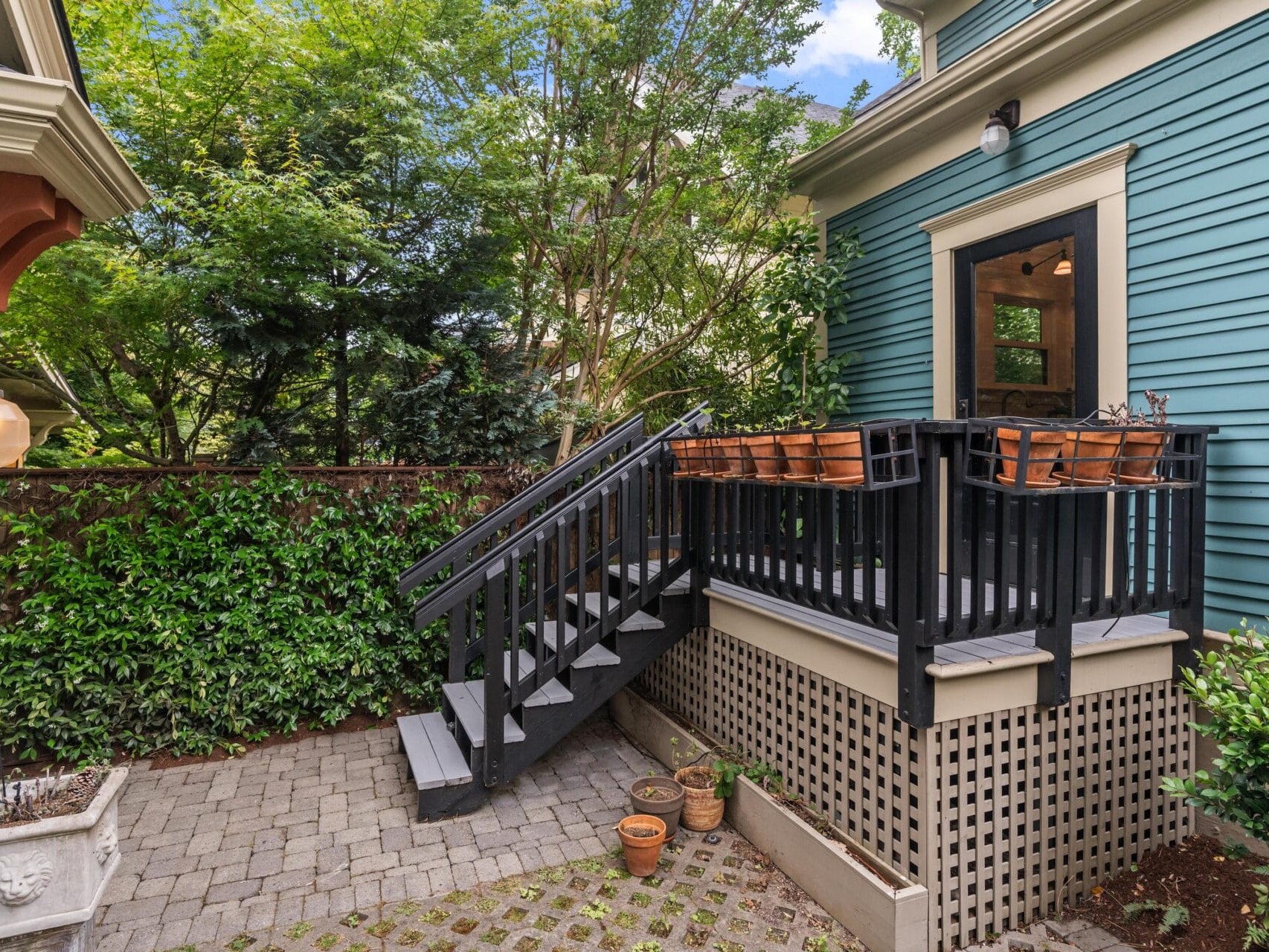 A small outdoor deck with black railings and wooden chairs, attached to a teal house. Stairs lead down to a paved patio surrounded by green hedges, trees, and potted plants.