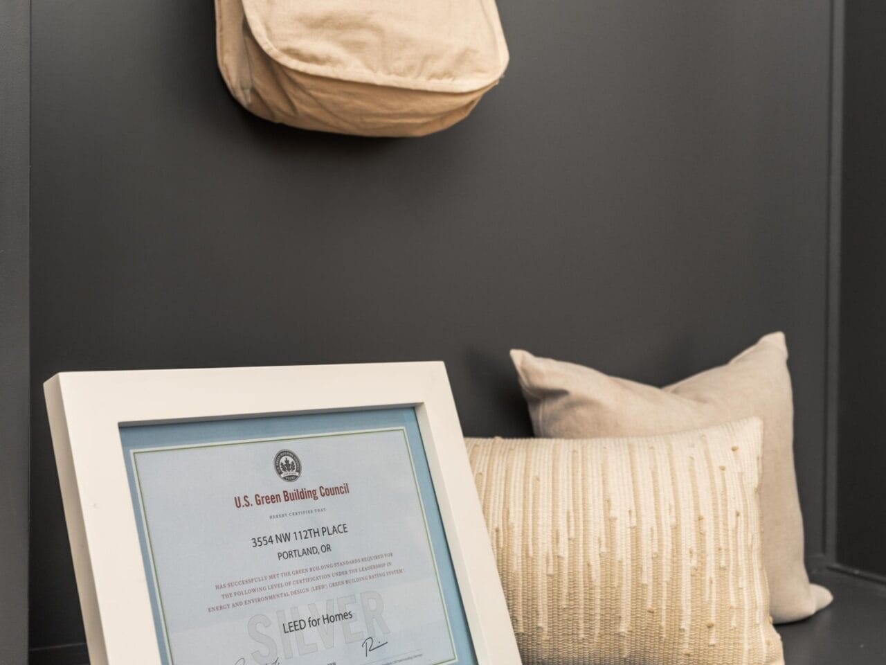 A framed certificate rests on a gray shelf next to cream-colored pillows. Above, a beige tote bag hangs from a hook on a dark gray wall.