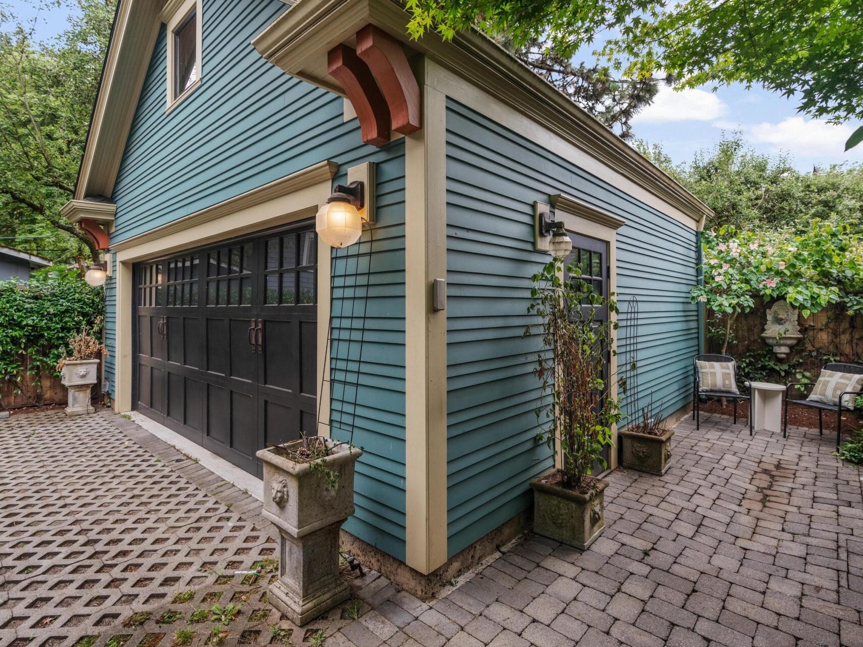 A blue garage with black doors sits at an angle, surrounded by a paved driveway and patio area with outdoor lighting, potted plants, and patio chairs under leafy trees.