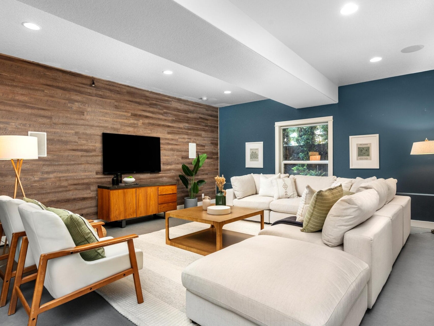 Modern living room with a large sectional sofa, two armchairs, wooden coffee table, and TV on a wooden accent wall. Blue and white walls, large window, and decorative lighting create a cozy, inviting space.
