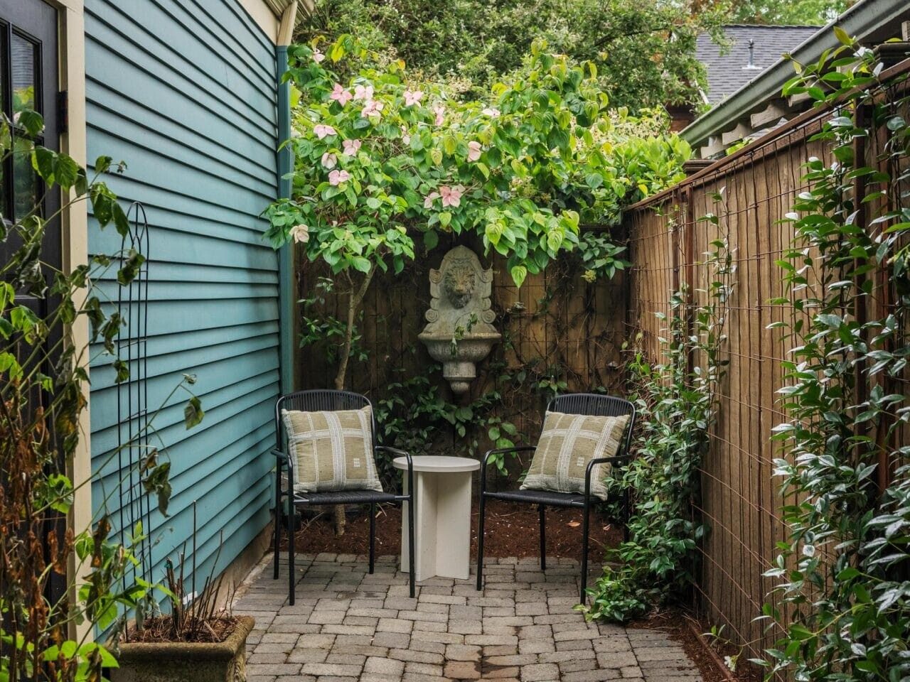 A small brick patio with two chairs, a small table, and a wall-mounted decorative fountain, surrounded by lush greenery and vines. The area is bordered by a blue house wall and a wooden fence.