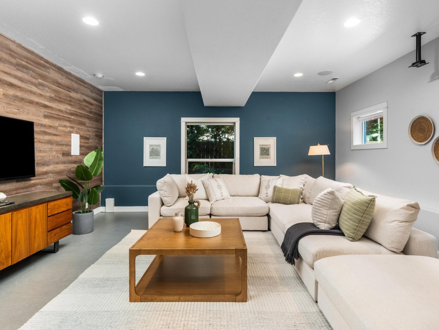 A modern living room with a sectional sofa, wood coffee table, TV on a wooden cabinet, potted plant, and wall art. The walls are blue and white with wood paneling, and a large window lets in natural light.