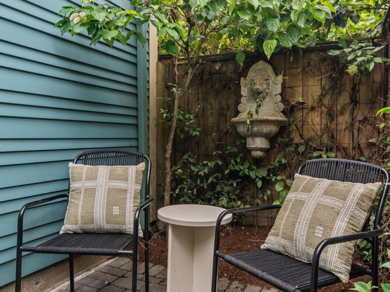 Two black chairs with patterned cushions face a small round table on a stone patio, bordered by greenery and a wooden fence with a decorative stone wall fountain.