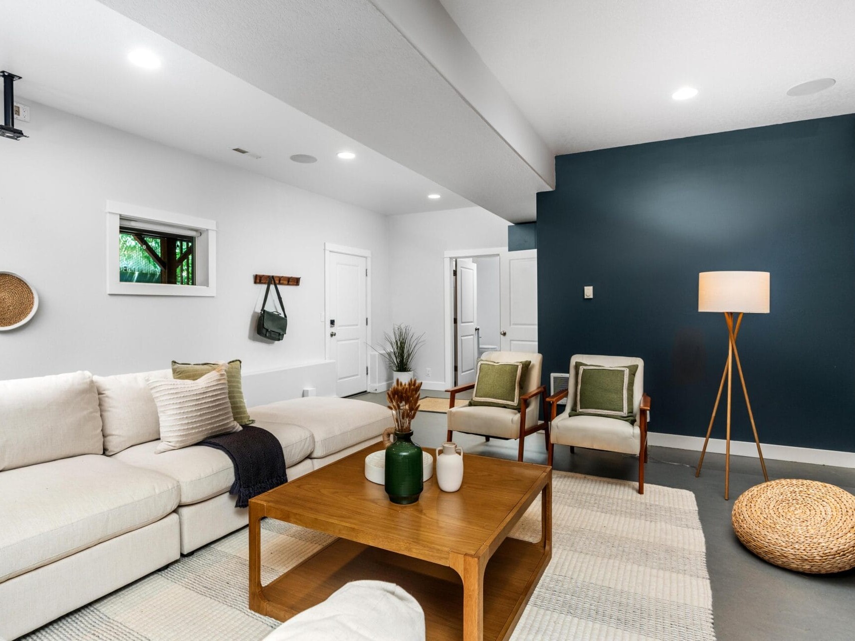 A modern living room with a white sectional sofa, wooden coffee table, two chairs with green cushions, a floor lamp, woven wall décor, and a dark accent wall. Natural light enters through a small window.