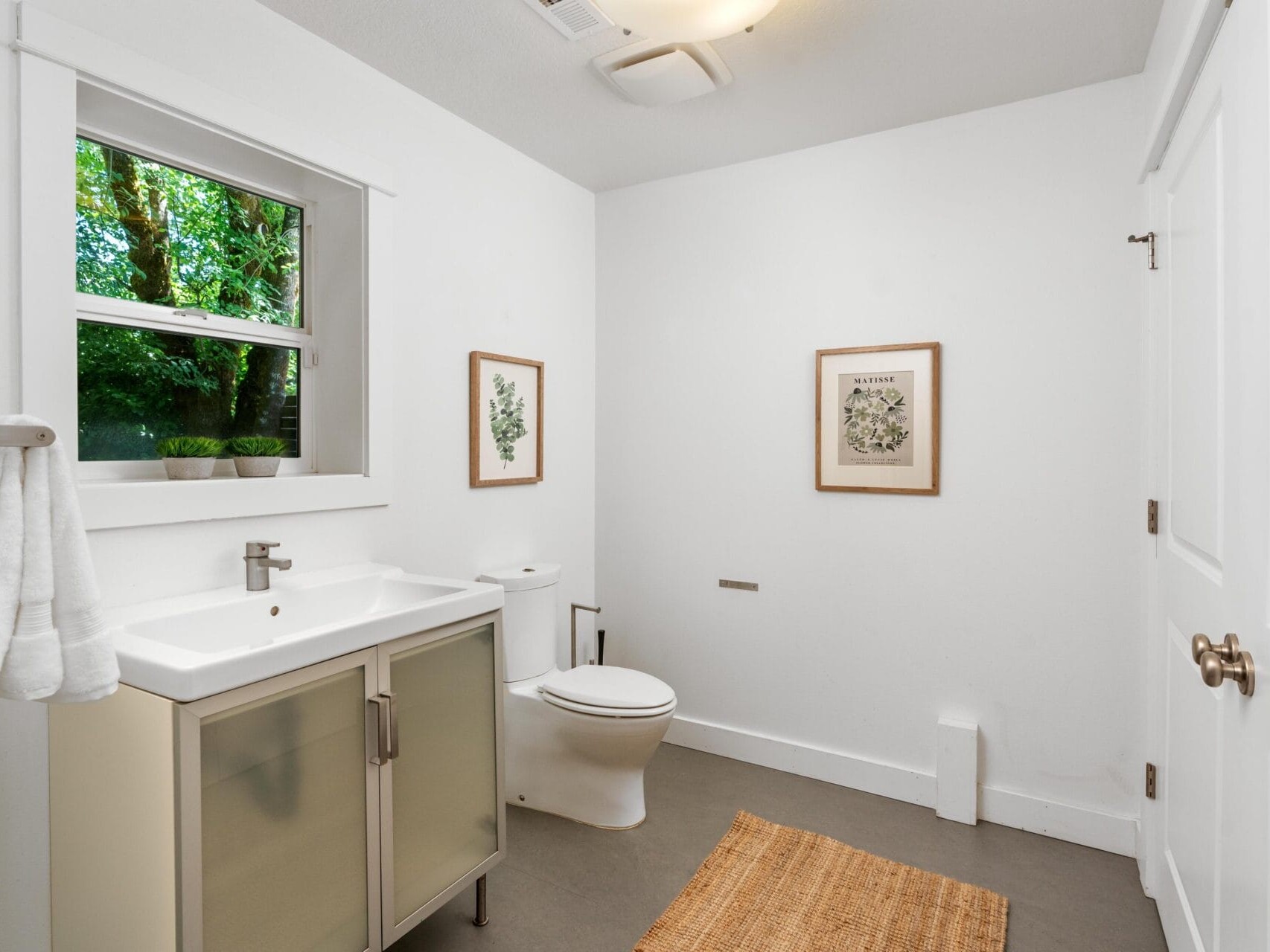 A modern, minimal bathroom with white walls, a single sink vanity, a toilet, two framed botanical prints, a window showing greenery outside, a woven rug, and a towel hanging by the sink.