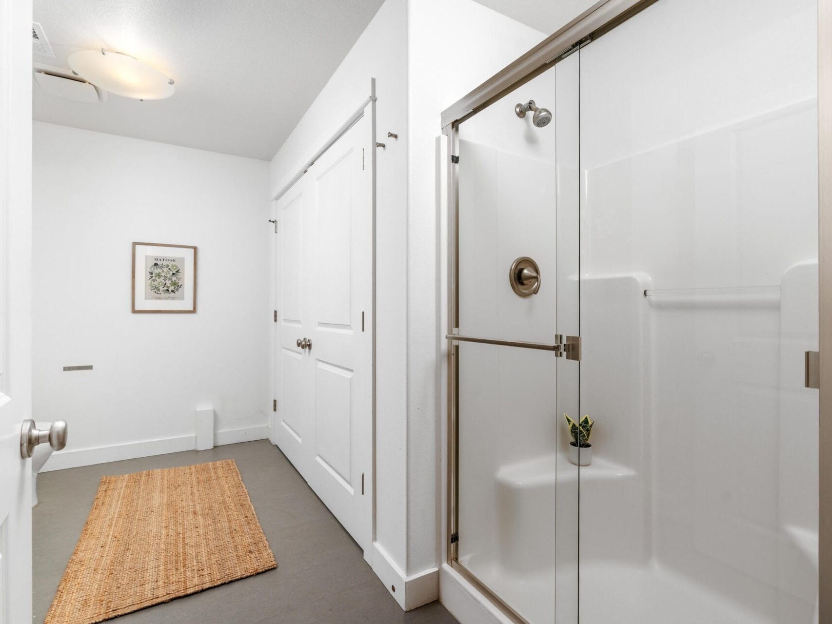 Modern bathroom with a glass shower enclosure on the right, double white doors in the center, a tan woven rug on the gray floor, white walls, and a framed picture hanging on the back wall.