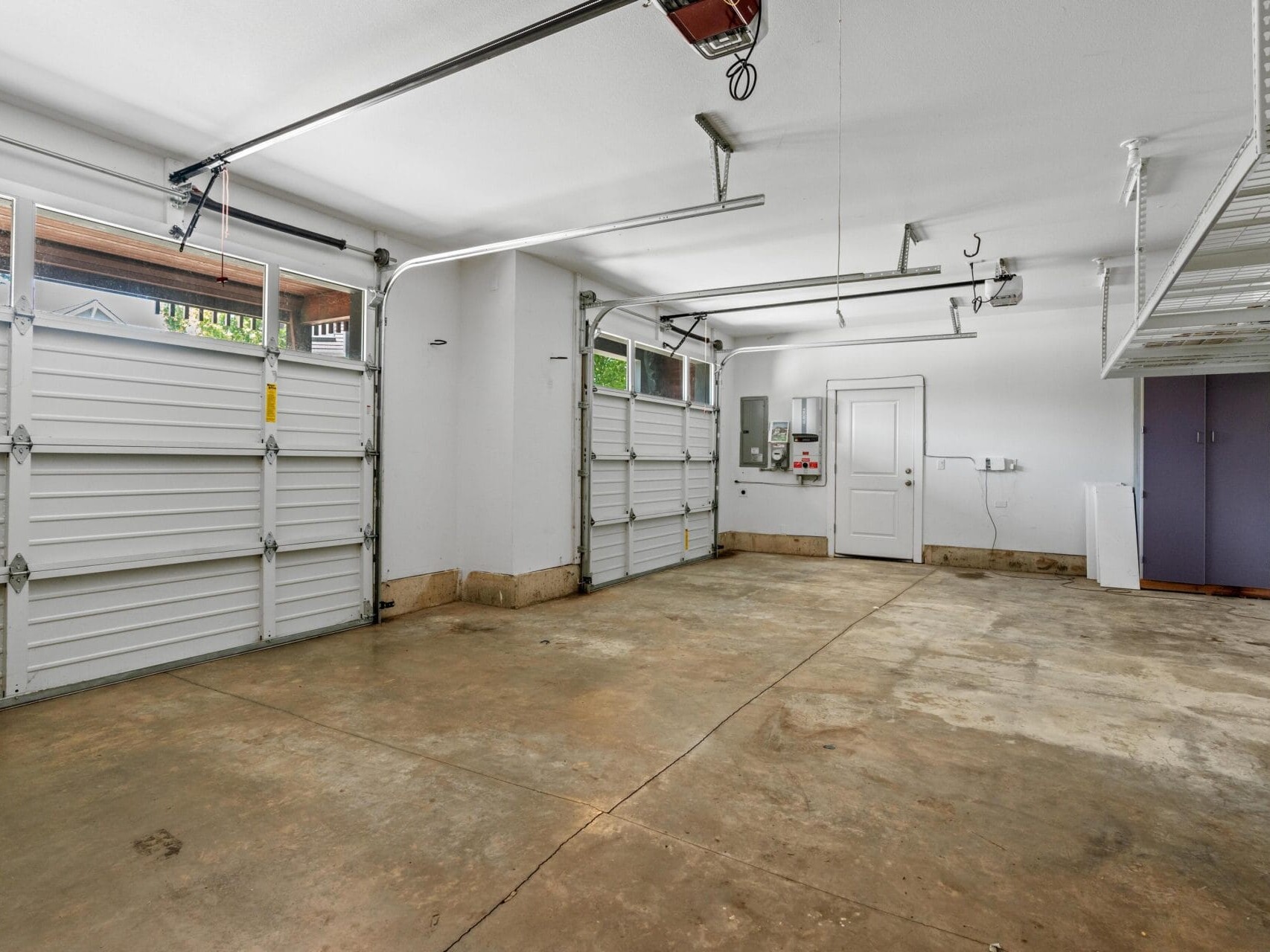 Spacious, clean two-car garage with concrete floors, white walls, two closed garage doors with windows, overhead storage racks, and utility panels on the back wall near a white door.