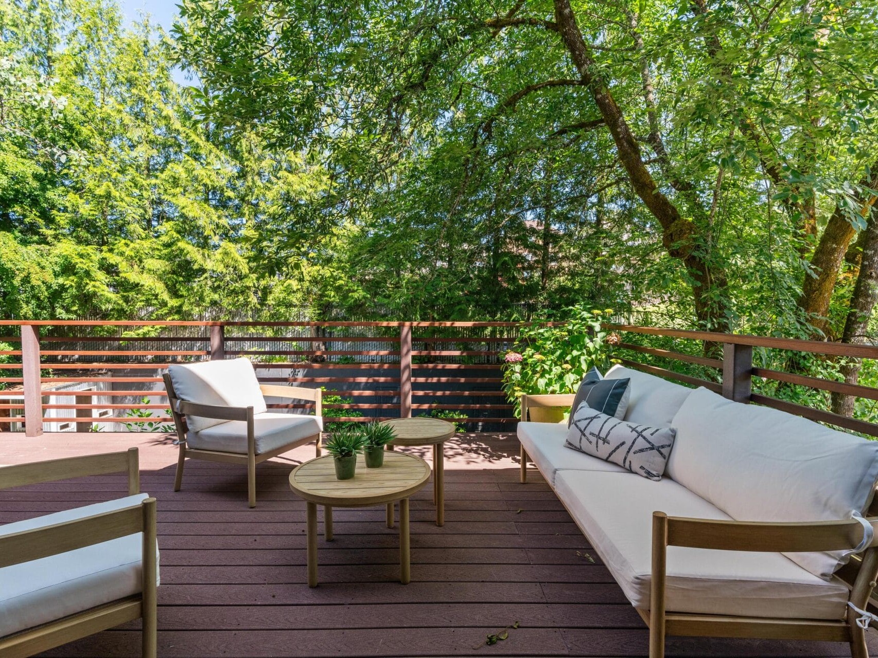 A wooden deck features cushioned patio furniture: a sofa, two chairs, and small round tables, surrounded by green trees and bathed in sunlight. Pillows and potted plants add a cozy touch.
