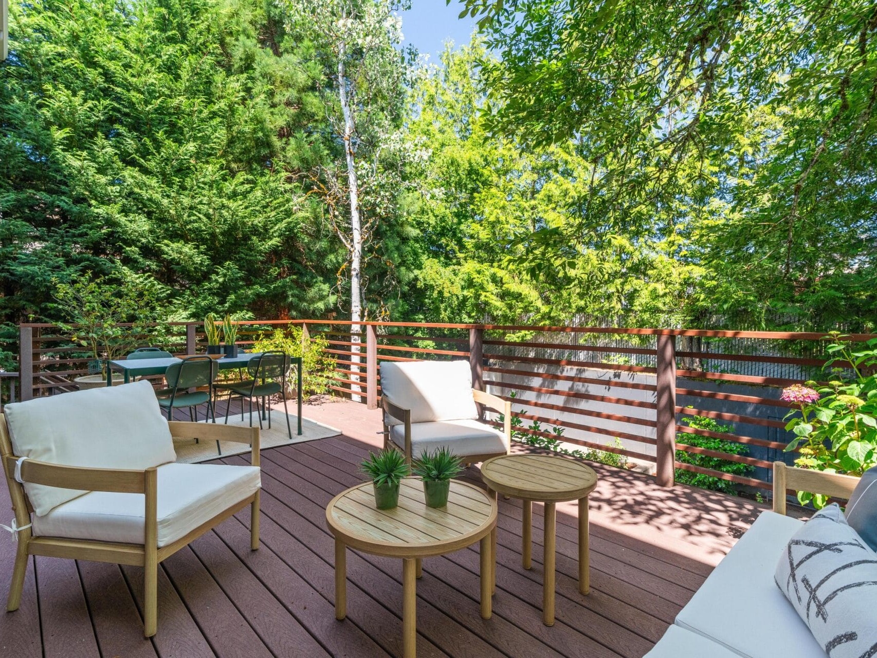 A wooden deck patio with cushioned chairs, a sofa, round coffee tables with potted plants, and a dining table, surrounded by lush green trees and bathed in sunlight.
