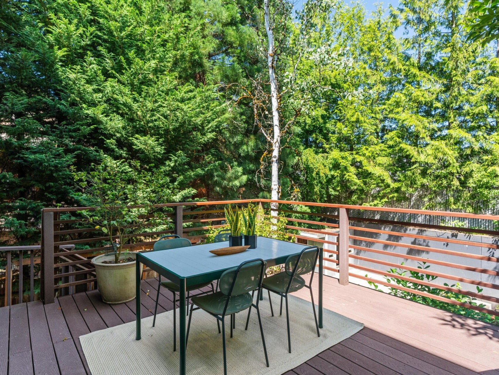 A modern outdoor deck with a blue table and four chairs on a beige rug, surrounded by potted plants and lush green trees, with sunlight filtering through the foliage.
