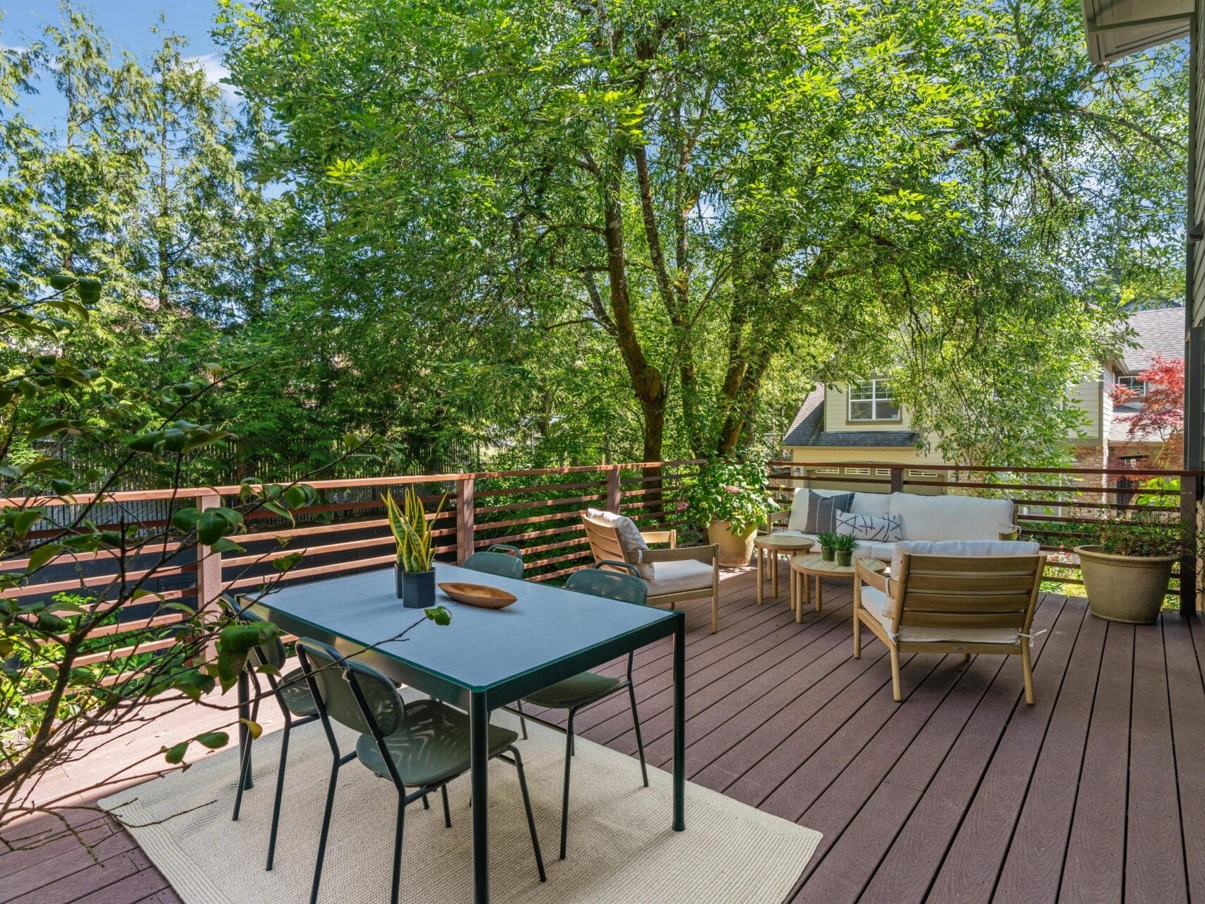 A spacious wooden deck with a dining table and chairs on a rug, a cozy seating area with armchairs and a sofa, surrounded by lush green trees and a wooden railing, attached to a house.